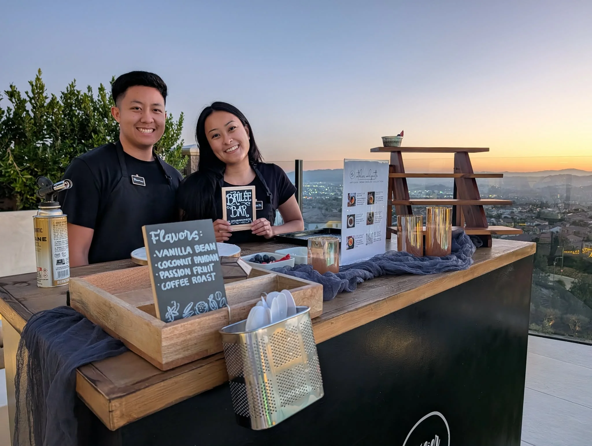 Two smiling people standing behind a coffee stand on a rooftop during sunset, with a cityscape in the background.
