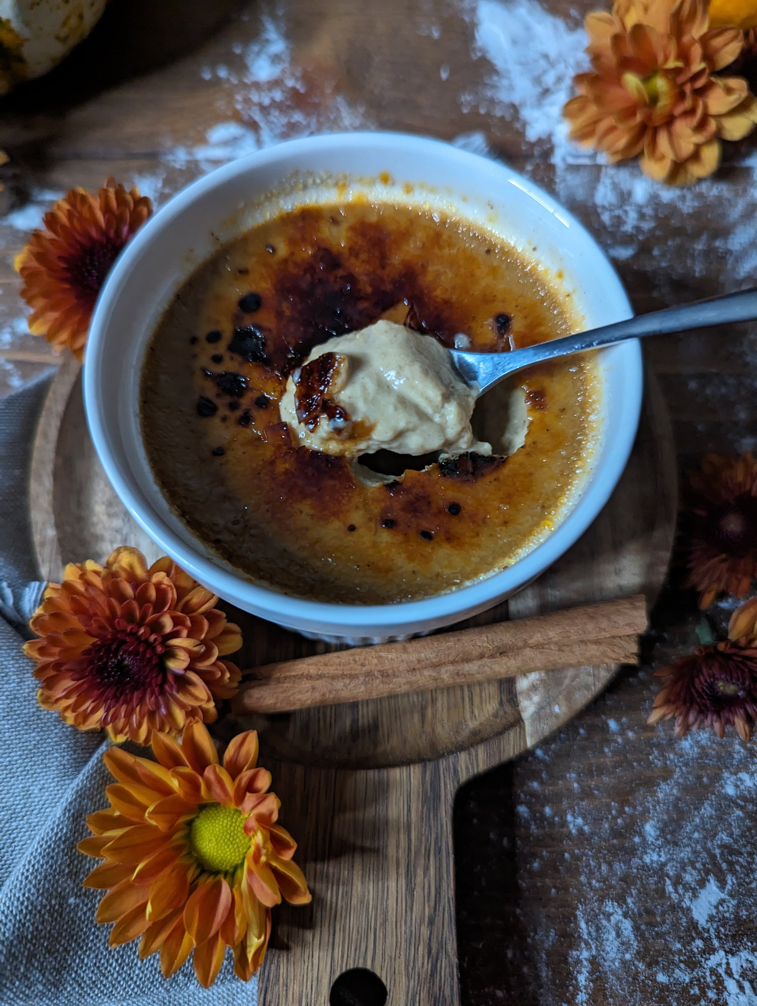 A ramekin of crème brûlée topped with a dollop of ice cream, surrounded by orange and red flowers on a wooden board.