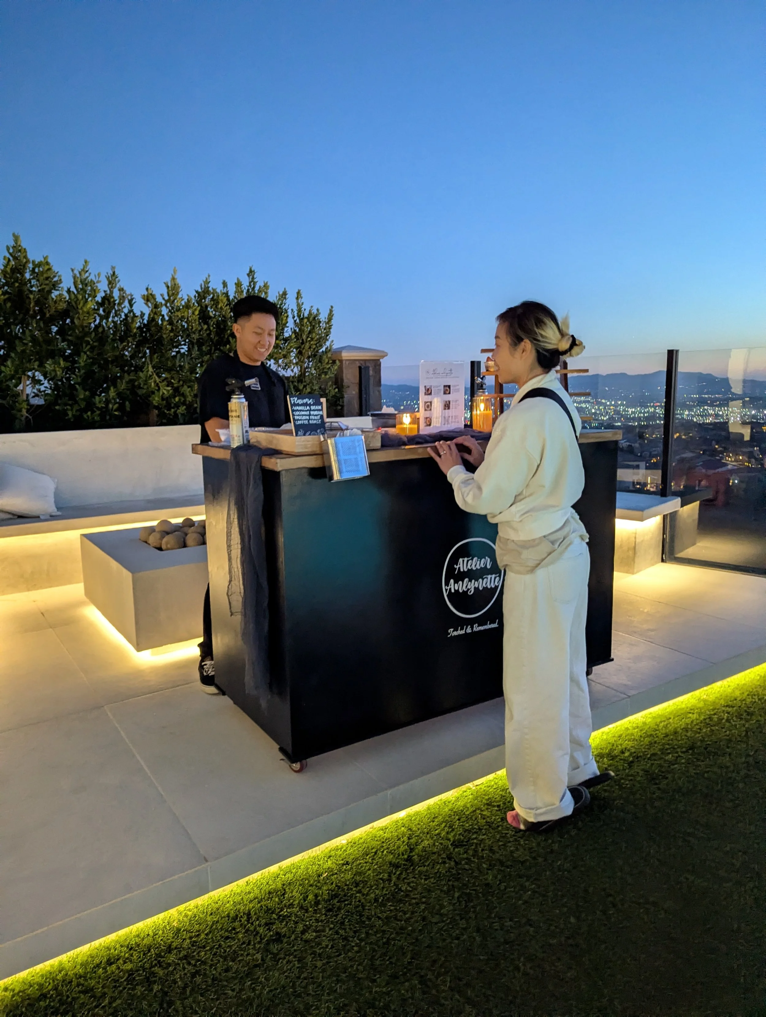 A woman in a beige jumpsuit standing at an outdoor rooftop bar during dusk, ordering drinks from a smiling bartender. The bar has a city view in the background with illuminated city lights, and the bar counter is decorated with candles and list displ
