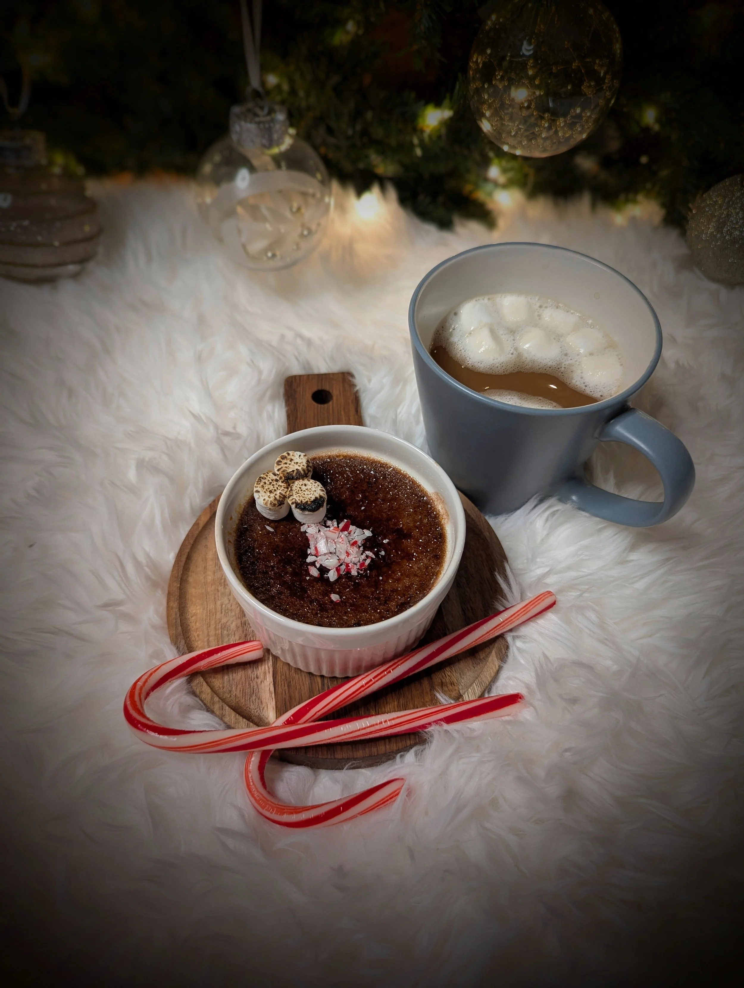 A cozy holiday scene with a cup of hot cocoa topped with mini marshmallows and marshmallow delights, a mug of coffee with cream, candy canes, and Christmas ornaments on a fluffy white surface under a decorated Christmas tree with lights.