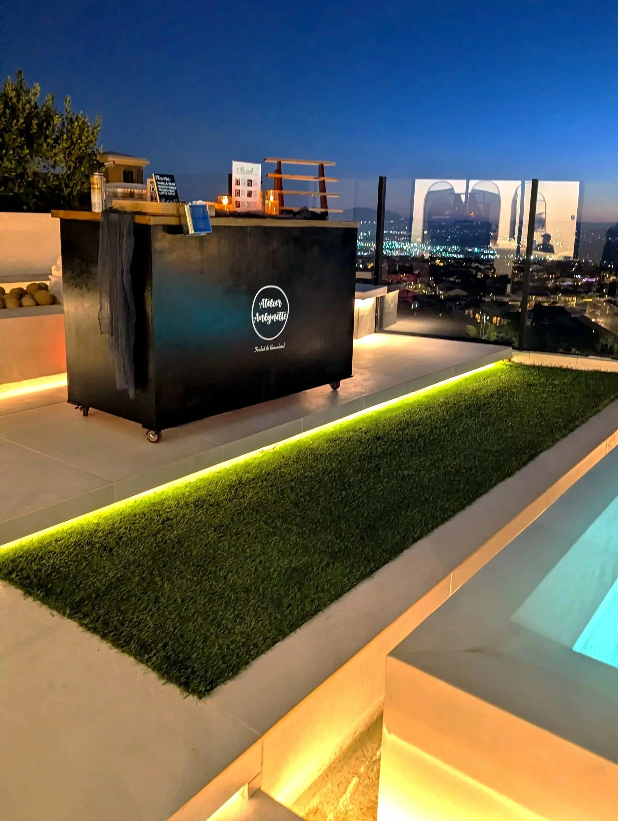 A rooftop bar or lounge with a city view at dusk. There is a black stand with a logo that reads 'Attnar Aromate' and some tables and decorations behind it. The area has ambient lighting, green grass inlay, and glass barriers overlooking the city skyl