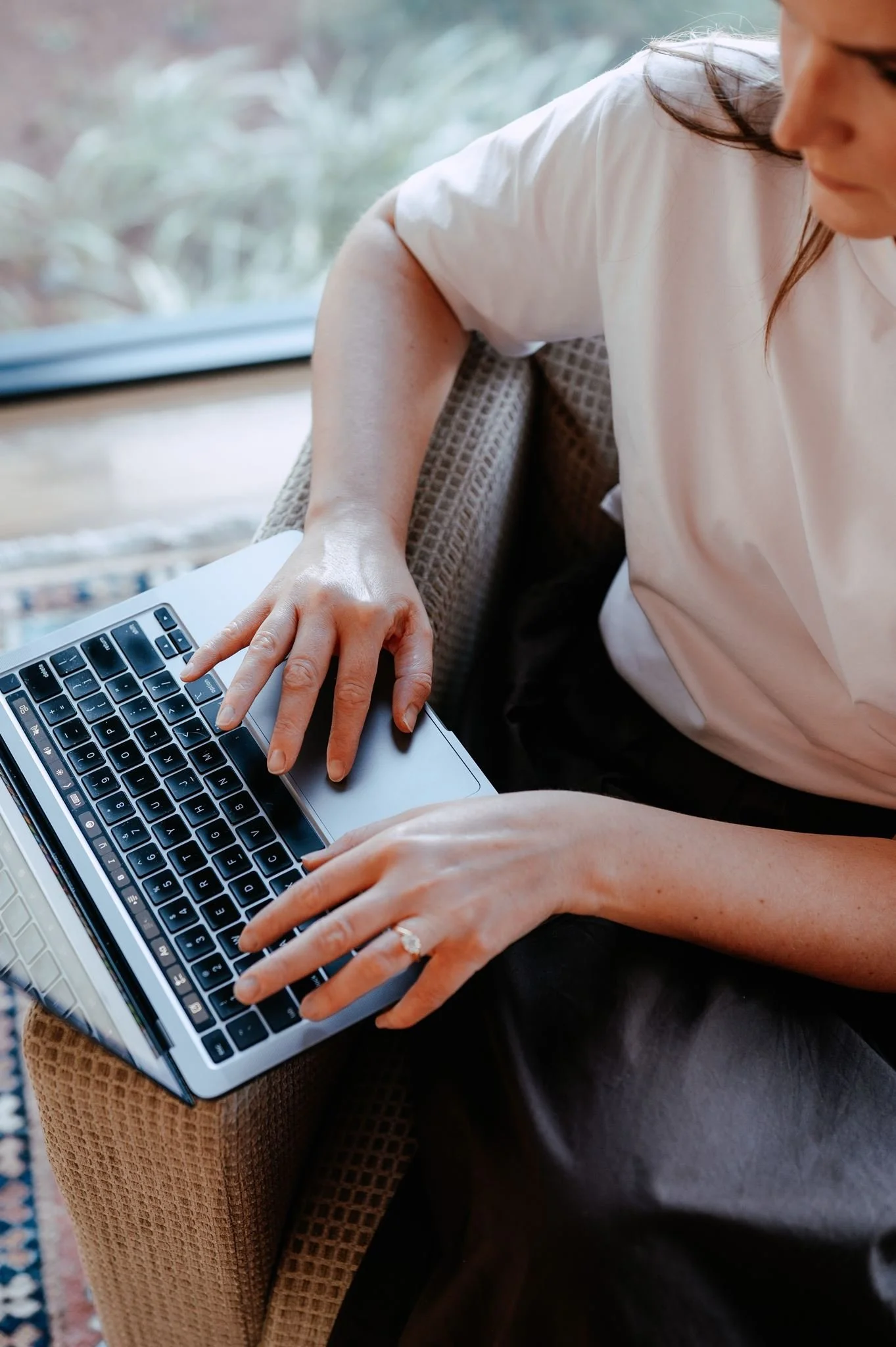 Close-up of a woman's hands typing on a MacBook laptop, sitting on a wicker chair with a colorful rug visible in the background.