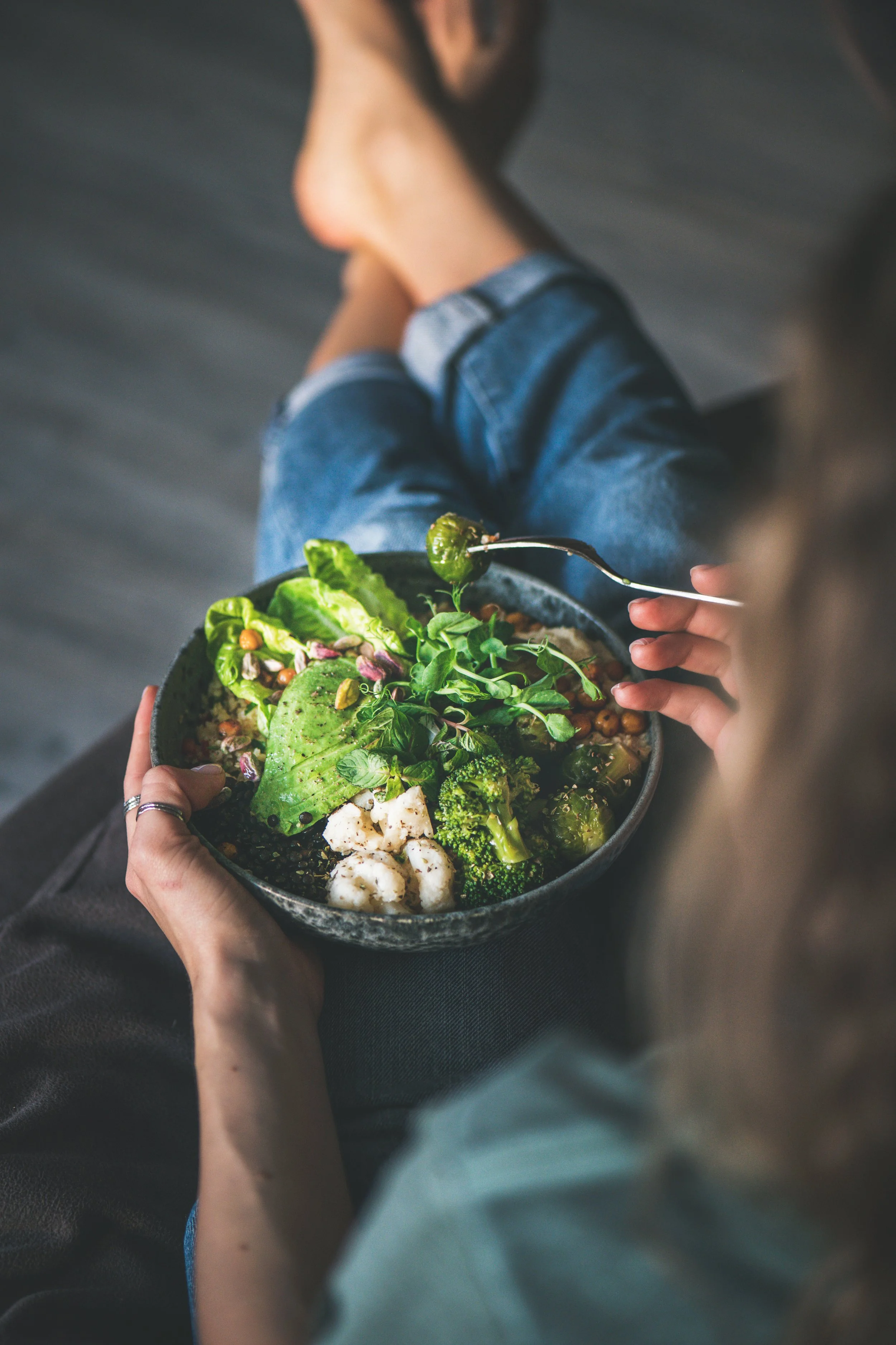 A person holding a bowl of mixed greens and vegetables, eating with a fork, with a background of a wooden floor and wearing jeans.