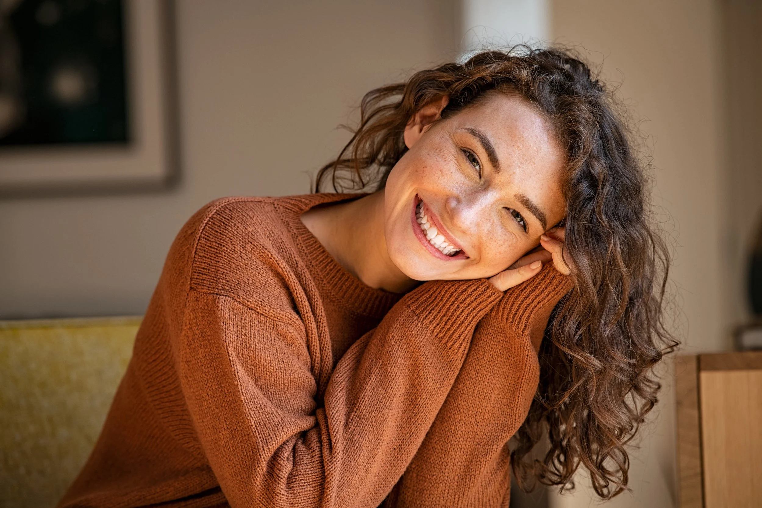 A woman with curly hair and freckles wearing an orange sweater, smiling while resting her head on her hands.