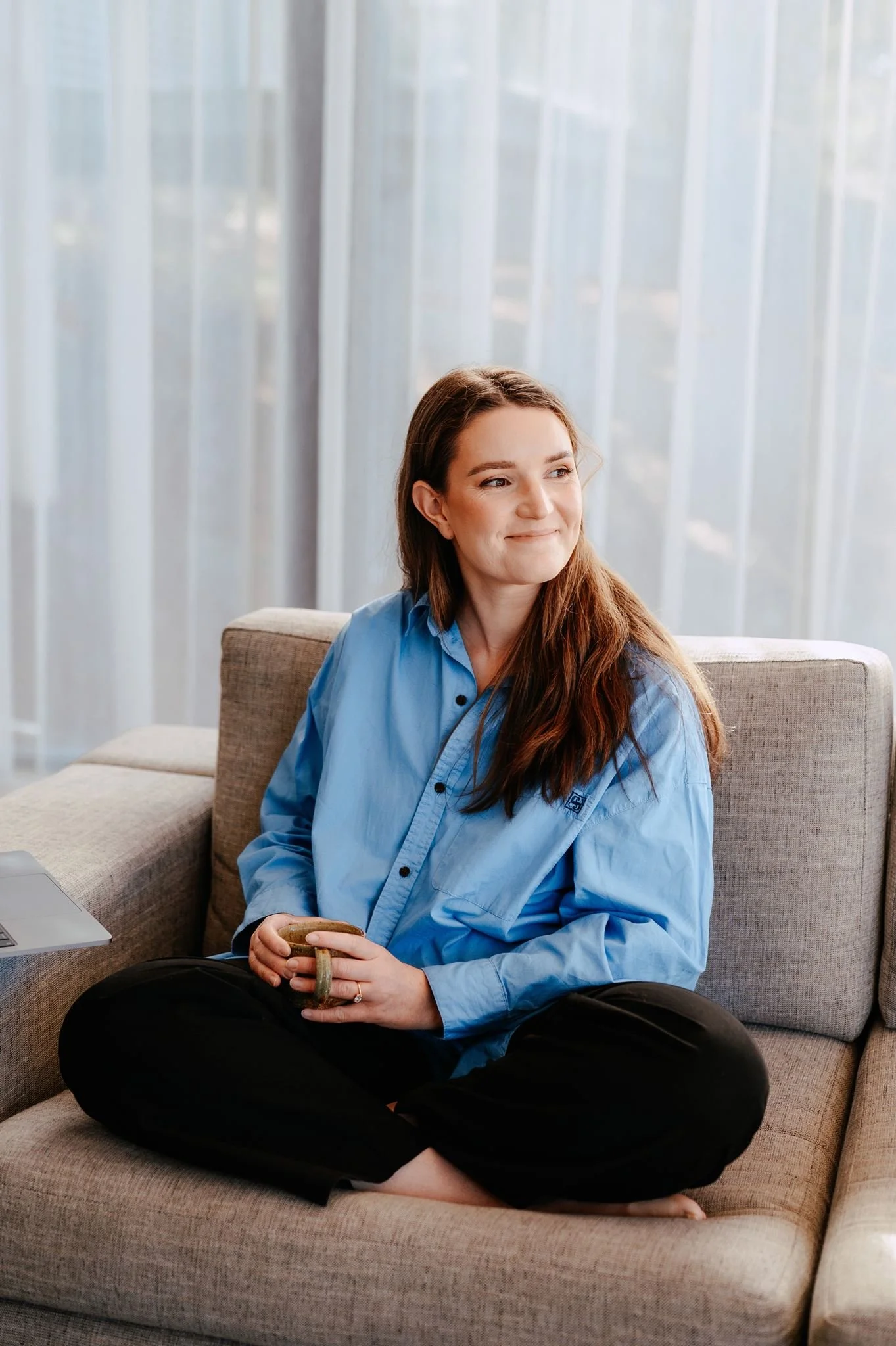 A woman with long brown hair sitting on a beige sofa, holding a mug, wearing a light blue shirt and black pants, smiling and looking to her left in a room with sheer curtains.