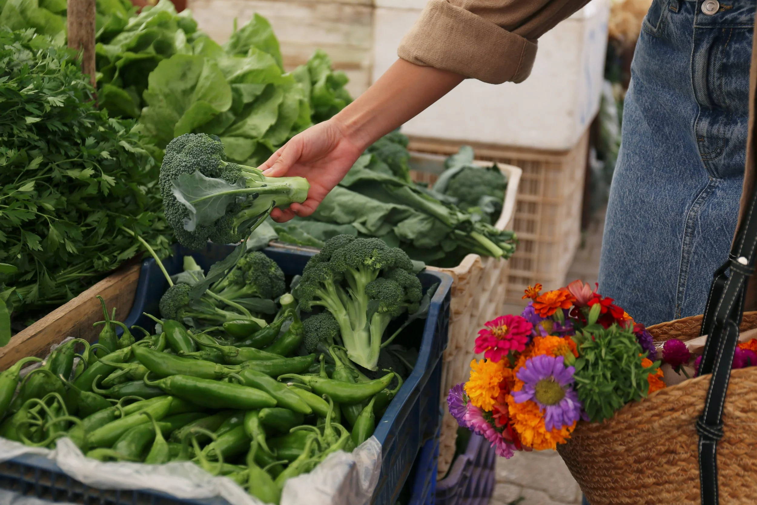 Person shopping for fresh vegetables at a farmers market, selecting broccoli, with baskets of colorful flowers and other vegetables visible.