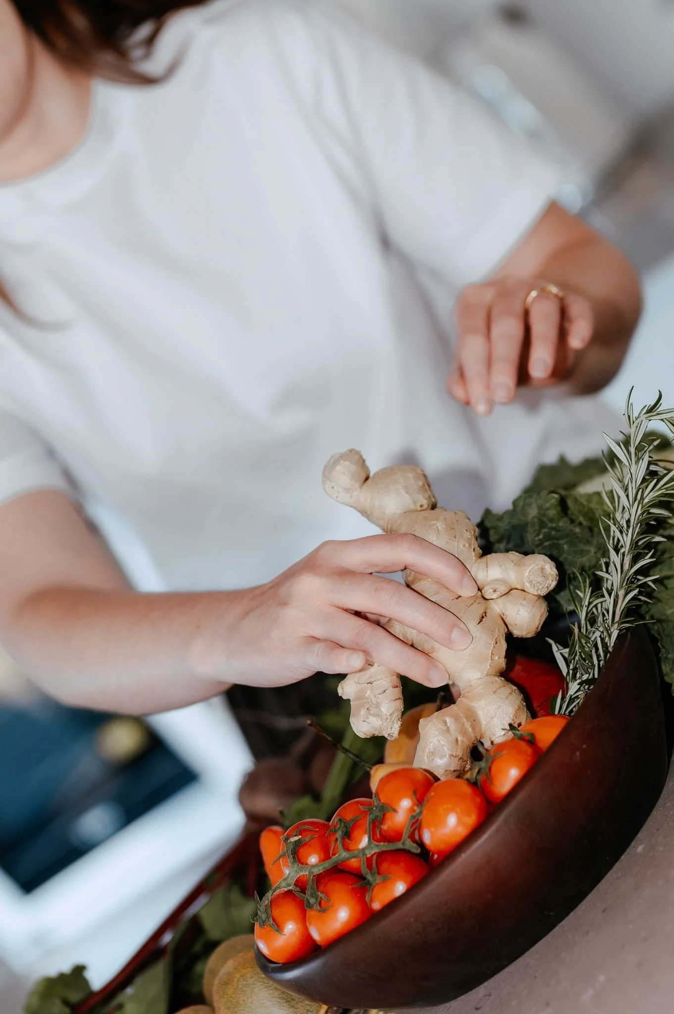 Person placing fresh ginger into a bowl of cherry tomatoes, surrounded by herbs and vegetables.
