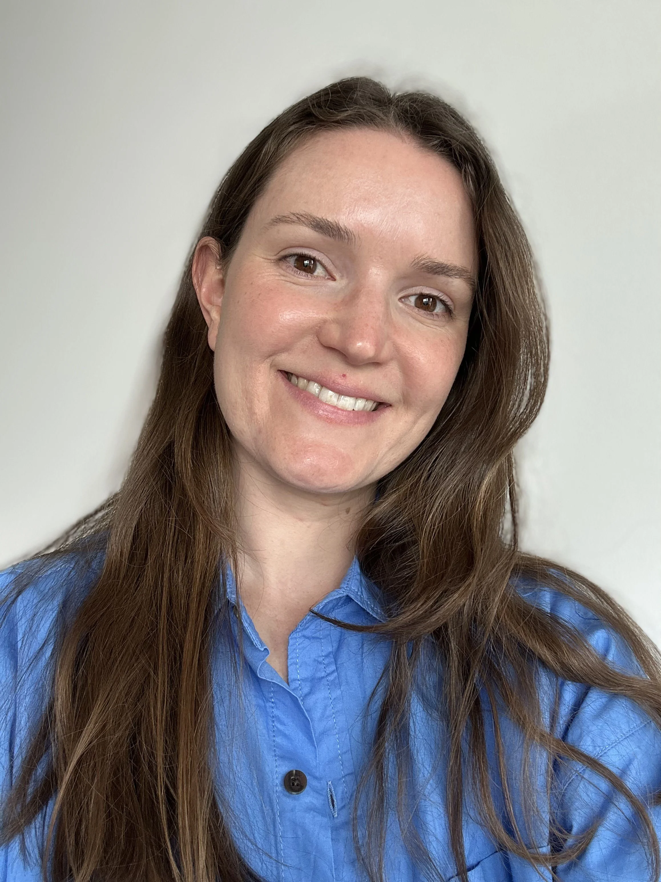 Close-up photo of a woman with long brown hair and light skin, smiling, wearing a blue button-up shirt, against a plain light background.