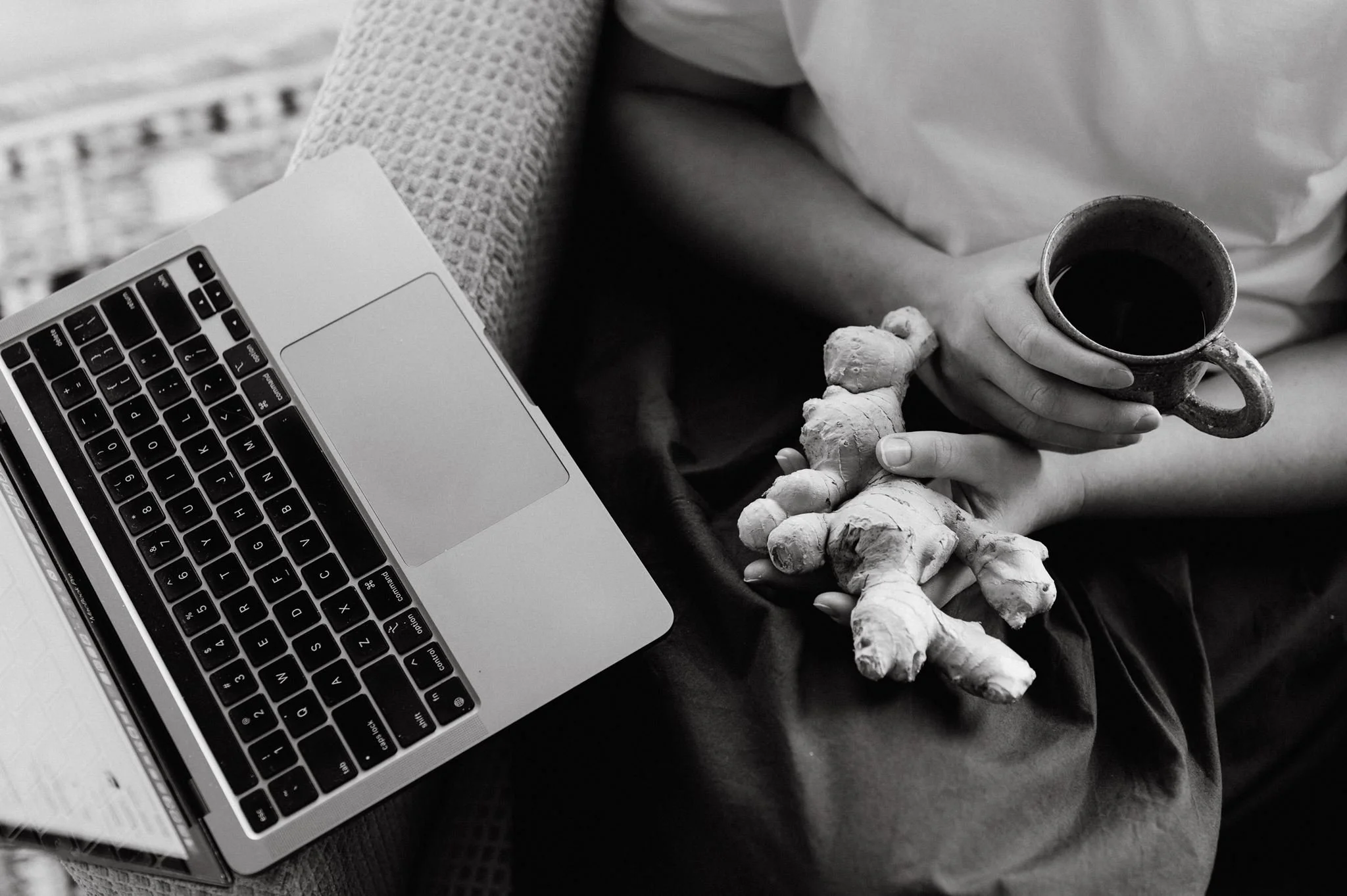 A person sitting with a laptop, holding a cup of coffee or tea, and a few pieces of ginger on their lap in black and white
