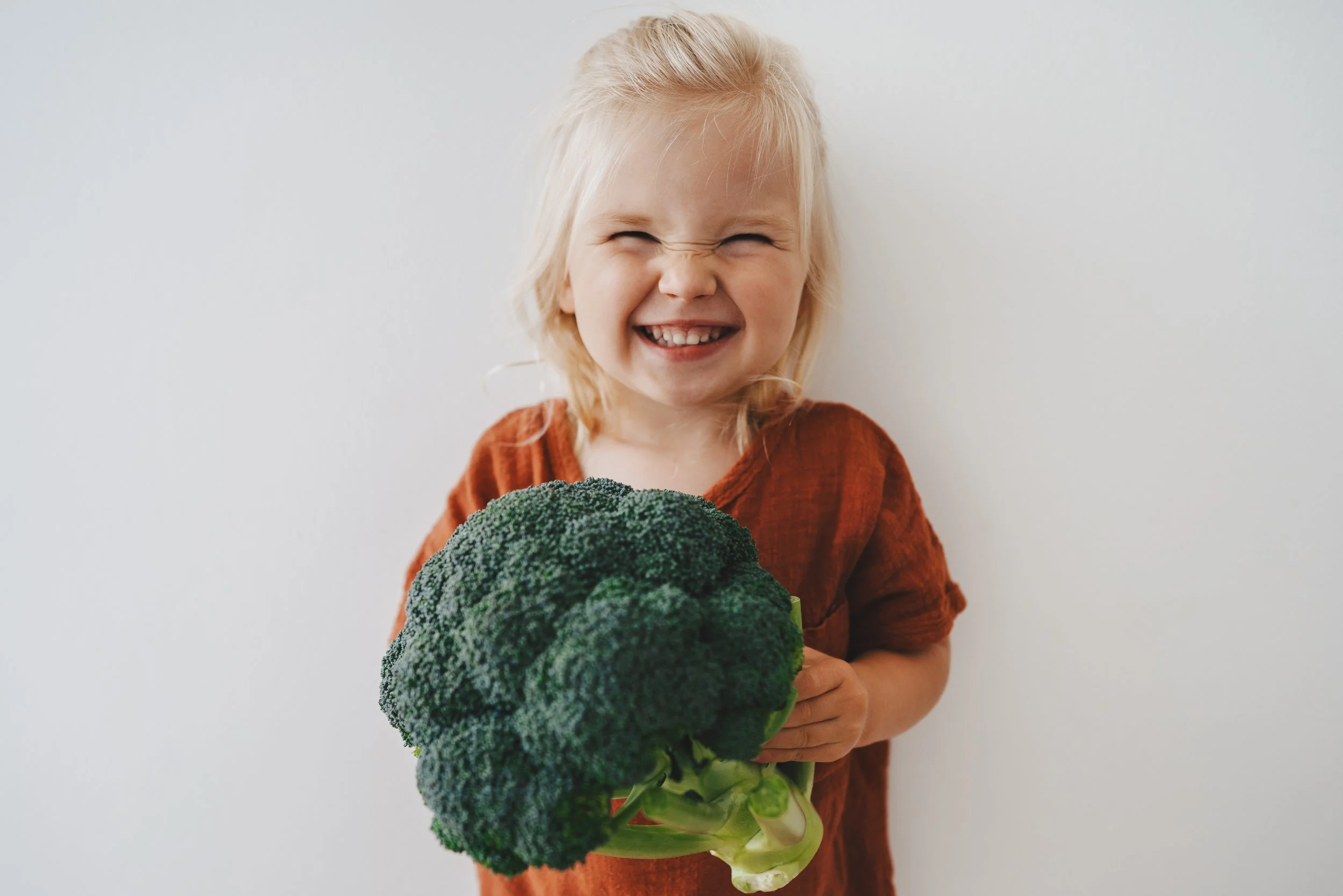 A smiling young girl with blonde hair holding a large head of broccoli.