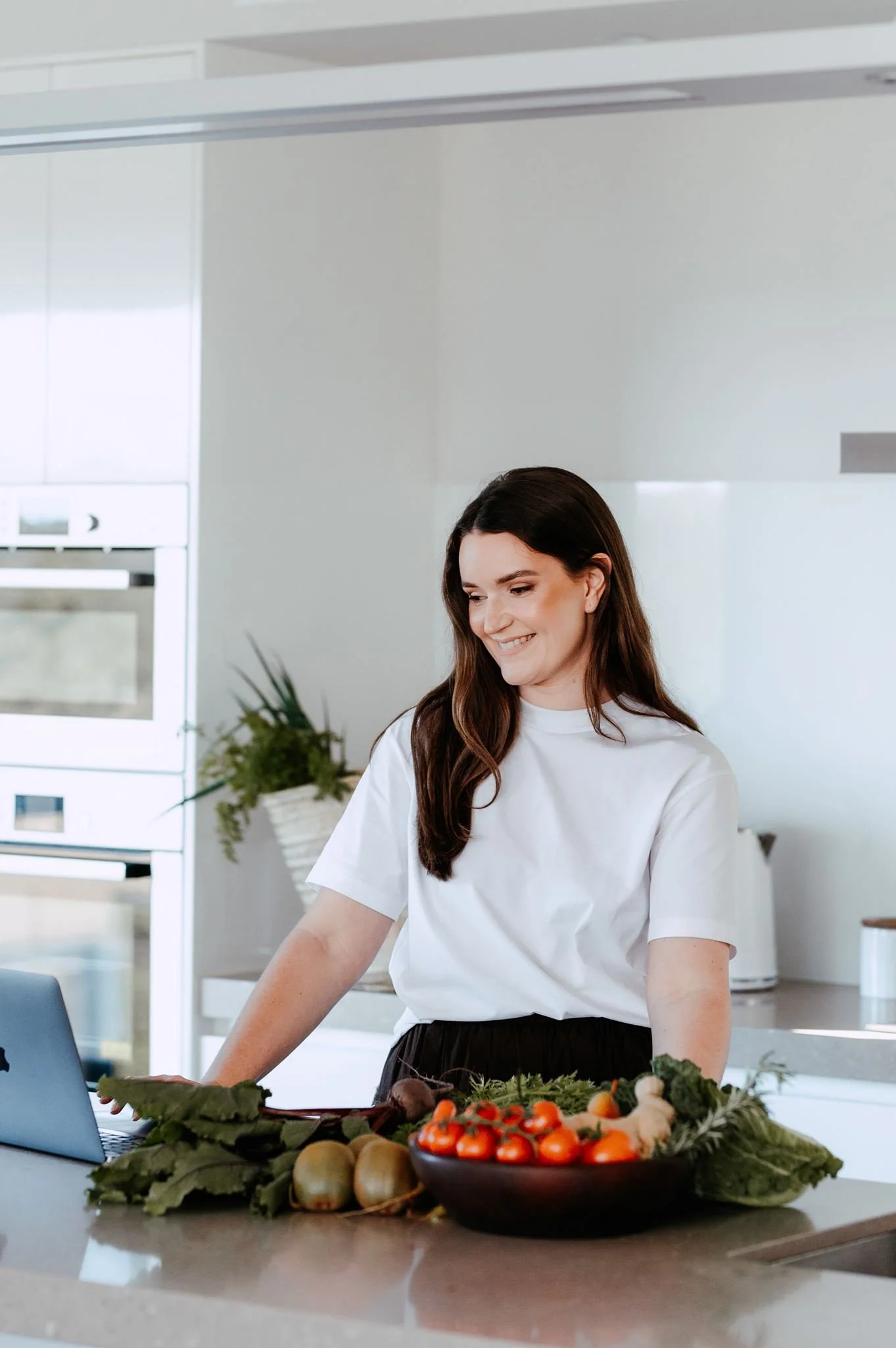 A woman with long brunette hair smiling while preparing vegetables at a kitchen counter with a bowl of cherry tomatoes, carrots, and other produce.