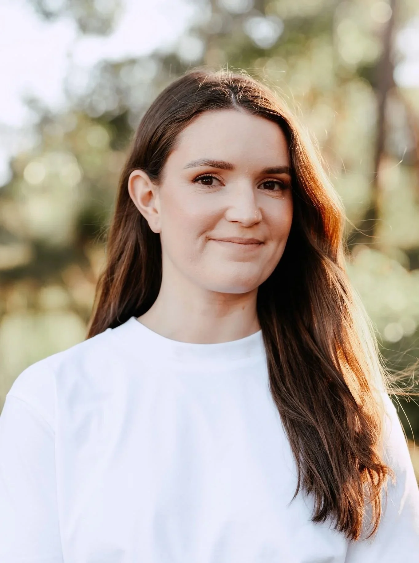 A young woman with long brown hair, wearing a white shirt, standing outdoors with blurred trees and sunlight in the background.