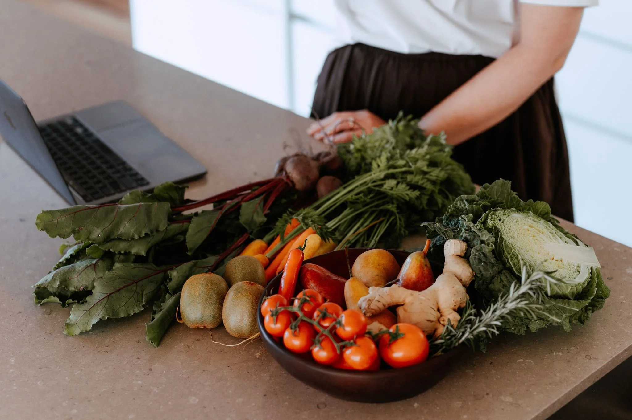 A person standing at a kitchen counter next to a laptop, with colorful fresh vegetables including carrots, tomatoes, cauliflower, leafy greens, and others in bowls and on the counter.