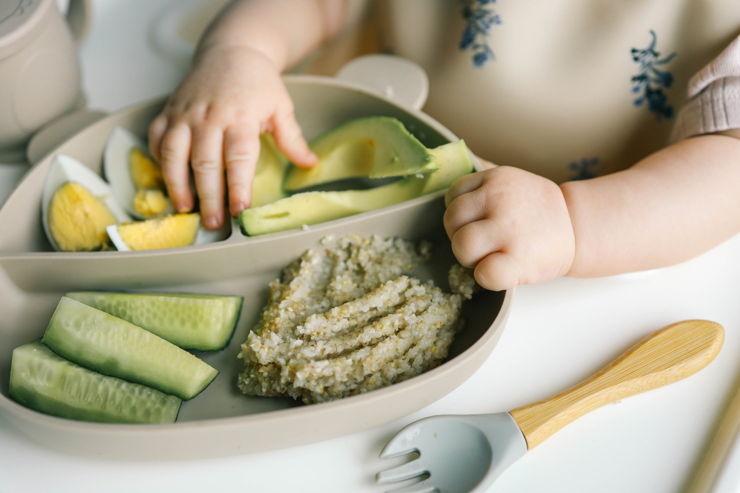 A child's hand reaching for a cucumber slice on a divided plate containing cucumber slices, hard-boiled egg quarters, and a serving of miso soup or porridge. An eating utensil with a wooden handle is on the white surface beside the plate.