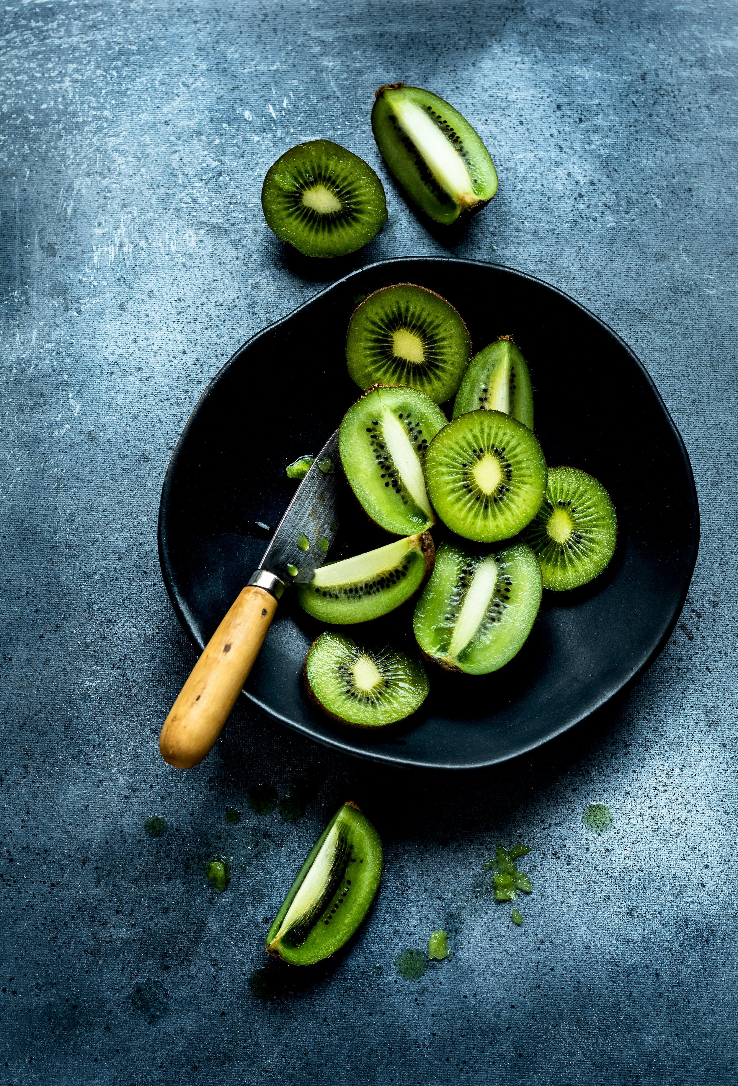 Sliced kiwi fruit in a black bowl with knife on a textured blue surface