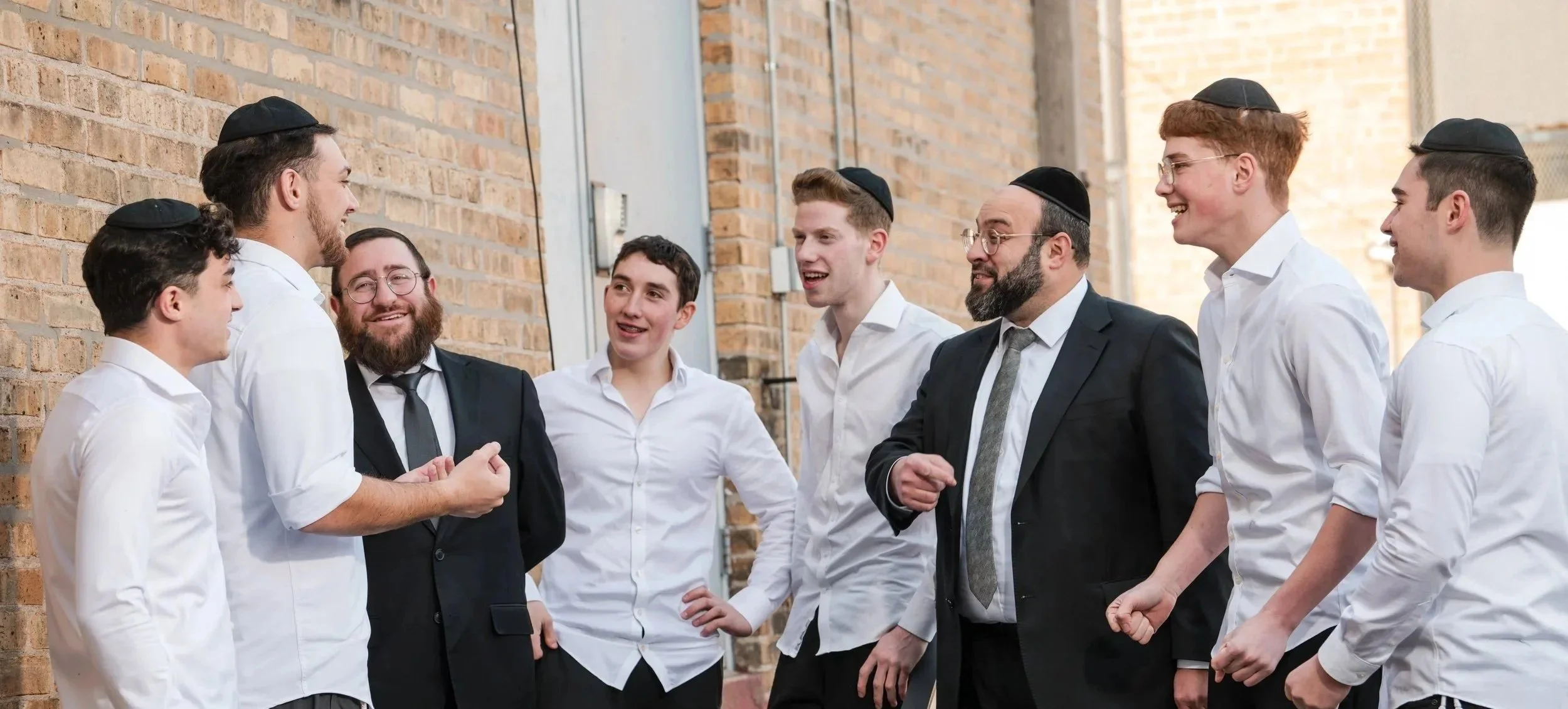A group of men wearing kippahs, some in religious attire, are laughing and talking outdoors against a brick wall during daytime.