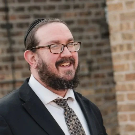 A smiling man with glasses, a beard, wearing a black suit, white shirt, and a patterned tie, standing outdoors in front of a brick wall.