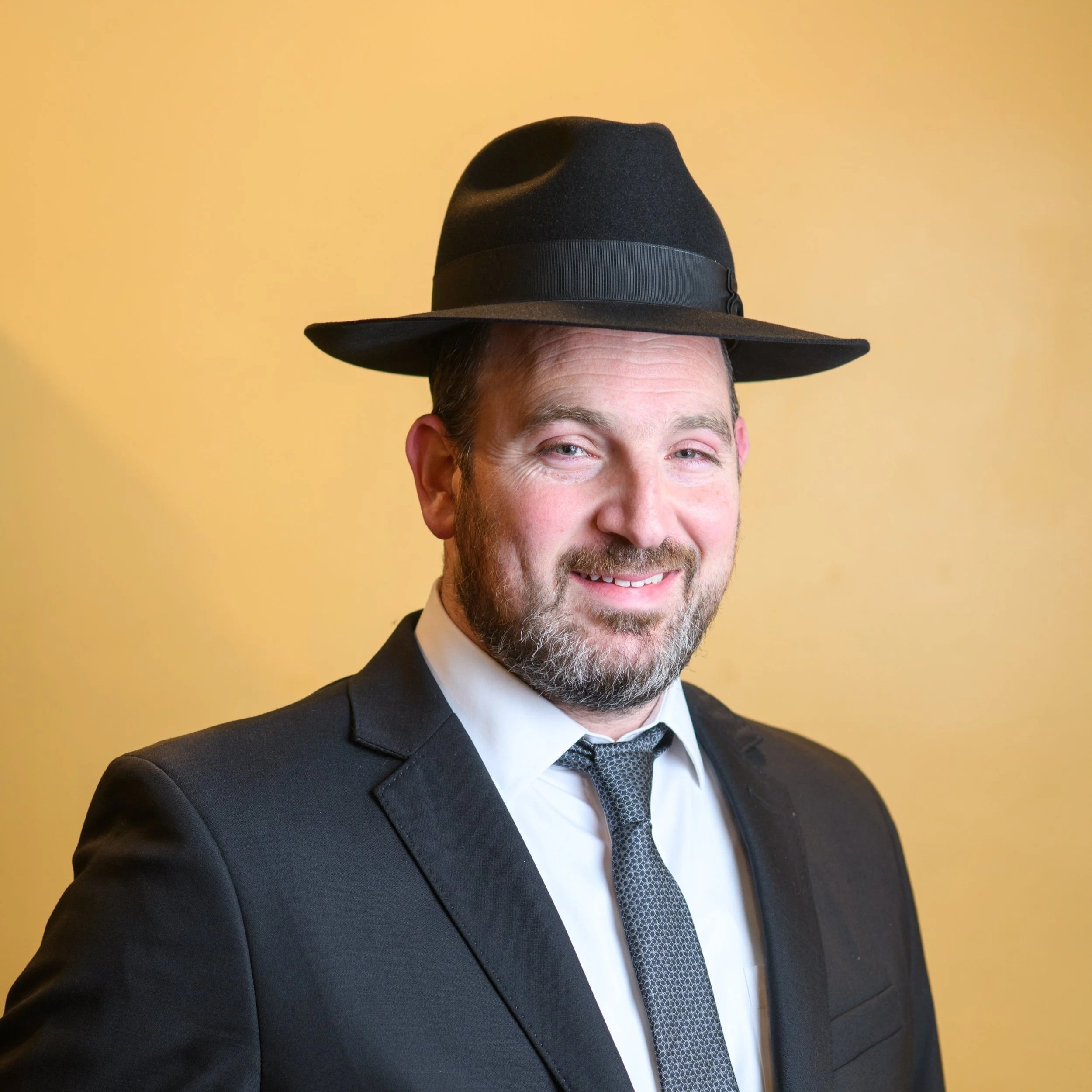 A man in a black suit with a white shirt and a patterned tie, wearing a black hat, smiling, with a yellow background.