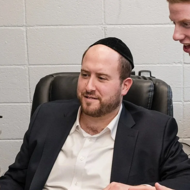 A man with a beard and a black kippah sitting in a black leather office chair, wearing a dark blazer and a white shirt, is looking down and smiling while another person to the right speaks to him.