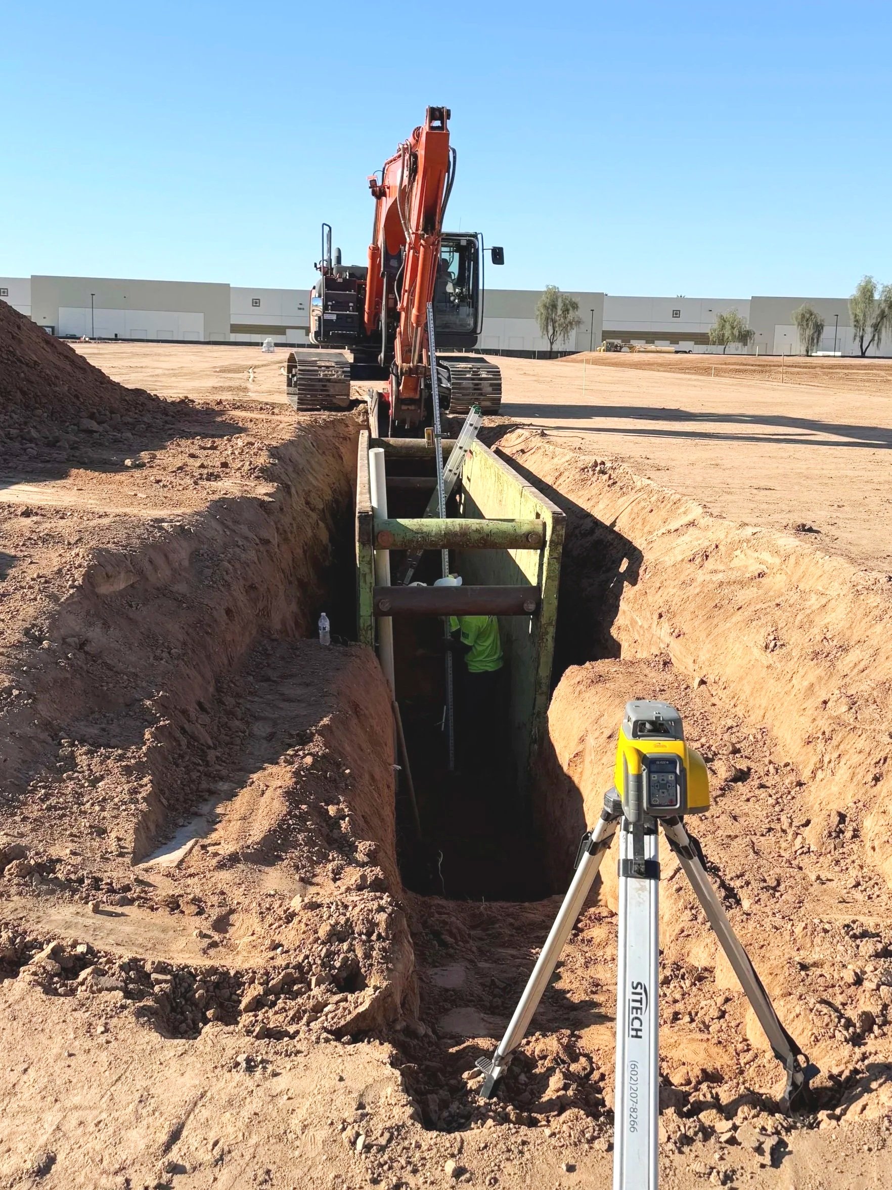 Construction site with a deep trench being excavated, a worker inside the trench, a yellow surveying instrument on a tripod in the foreground. GPS Equipment. Mesa, Phoenix, Queen Creek, Gilbert, San Tan Valley
