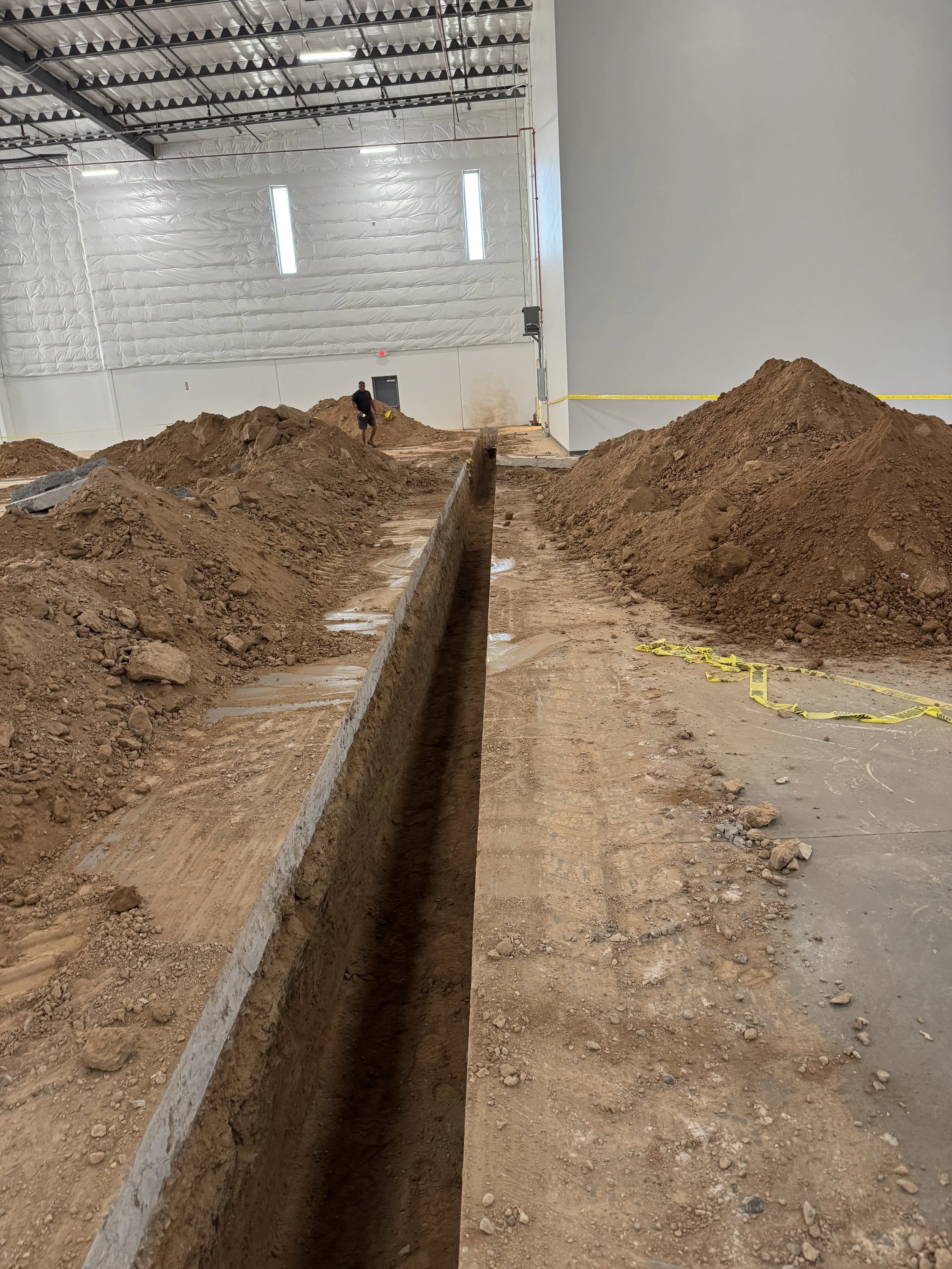Indoor construction site with a long trench in the ground surrounded by piles of dirt, with construction personnel working in the background, and yellow caution tape marking off the area.