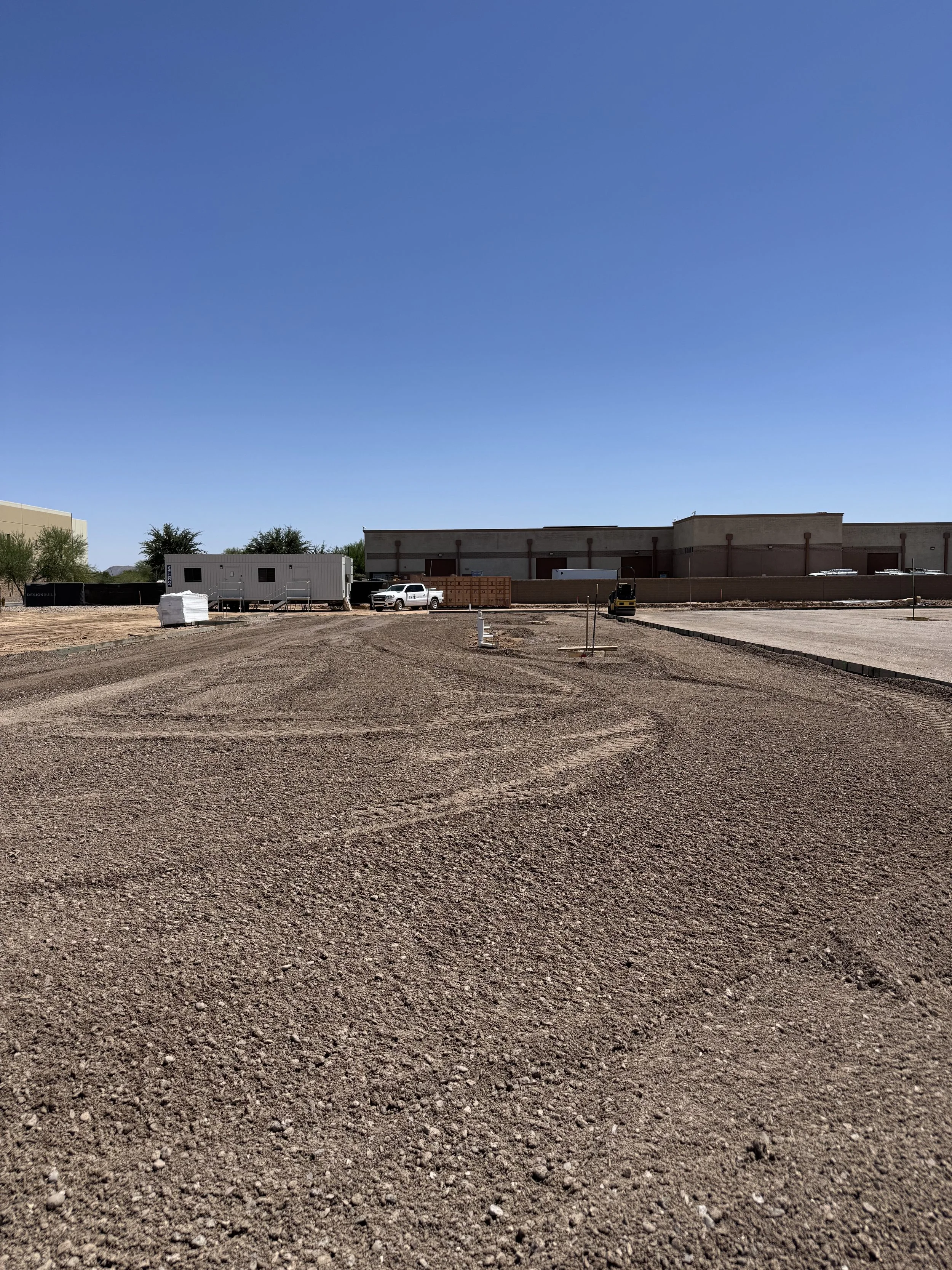 Construction site with dirt ground, construction equipment, and a partially paved area under a clear blue sky.