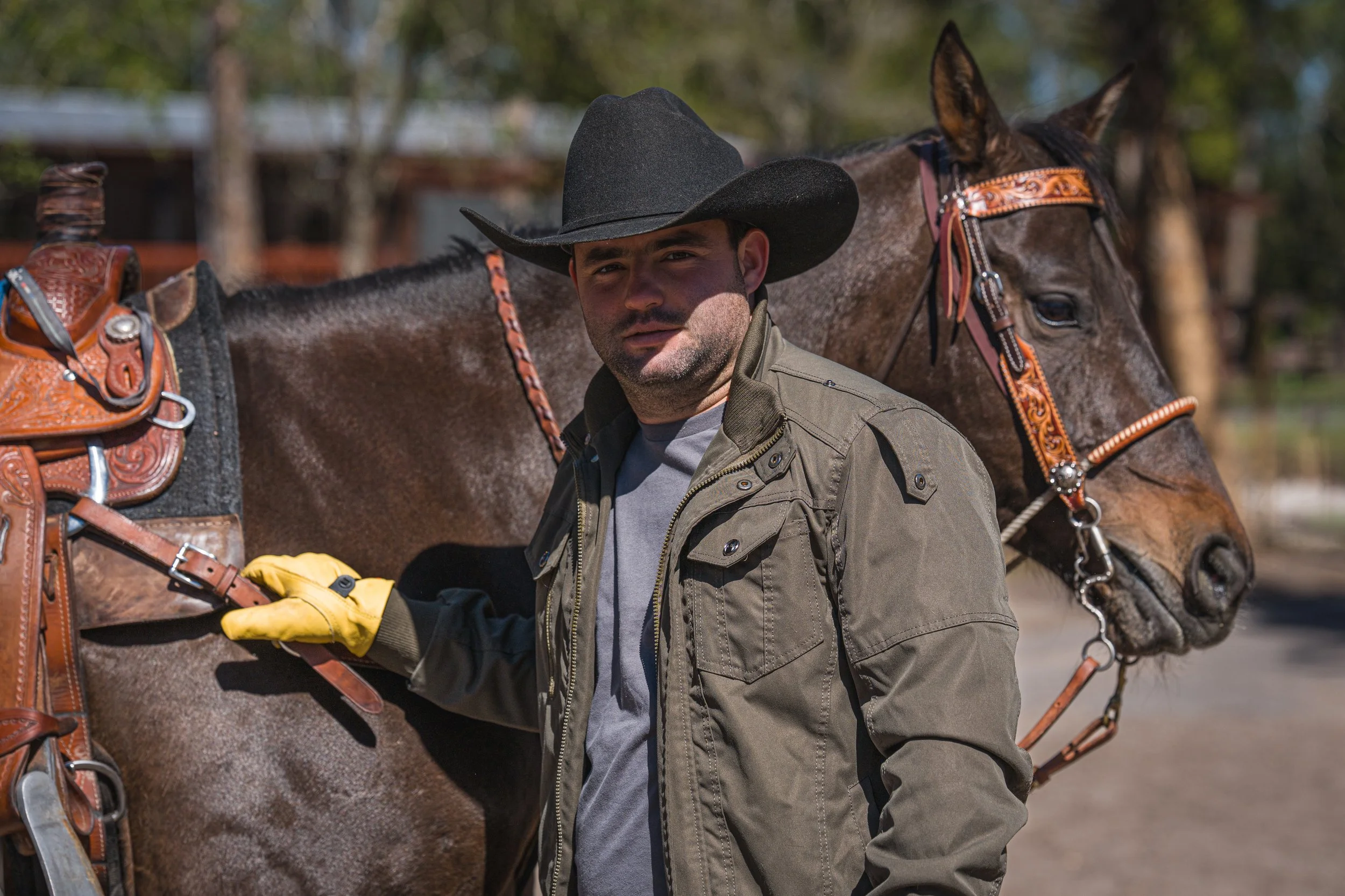 Hombre con sombrero de vaquero y chaqueta de camuflaje junto a un caballo con arnés decorado. Vaqueros Del Pantano.