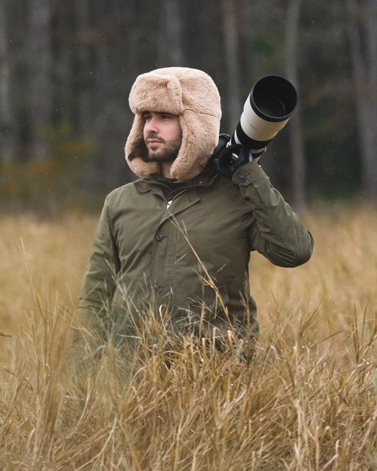 Eric Pneuma con abrigo verde y gorro de piel, sosteniendo un teleobjetivo grande, en un campo de hierba seca. Vaqueros Del Pantano.