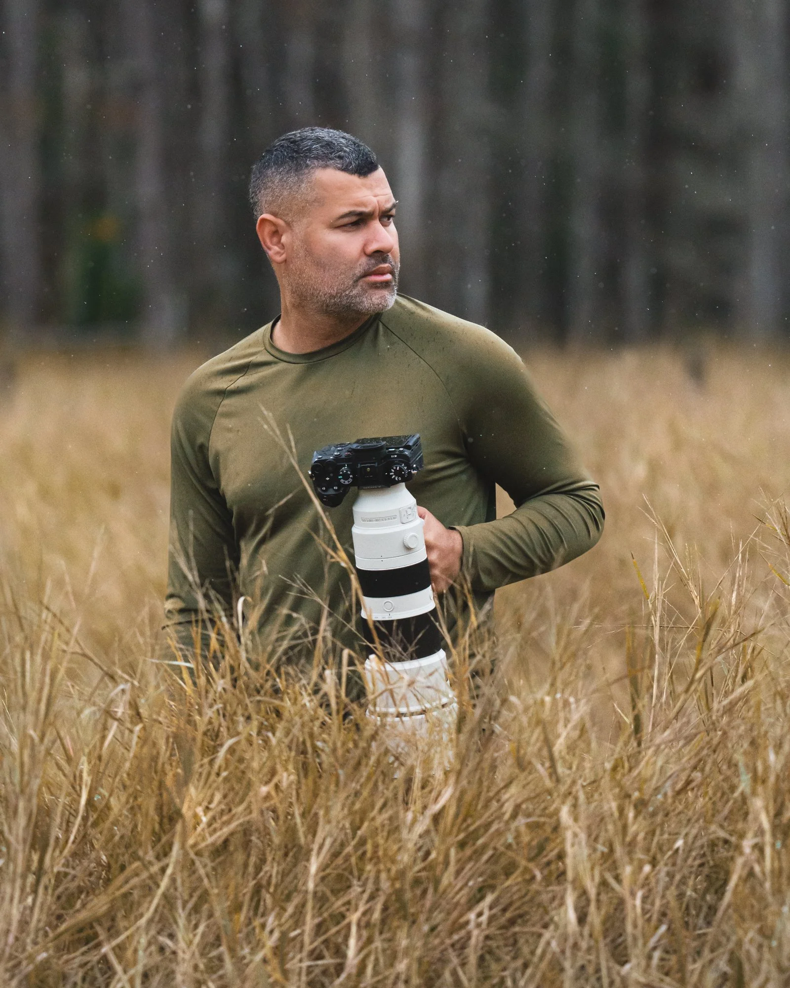 Gutti en un campo con vegetación, sosteniendo una cámara con un teleobjetivo grande. Vaqueros Del Pantano.