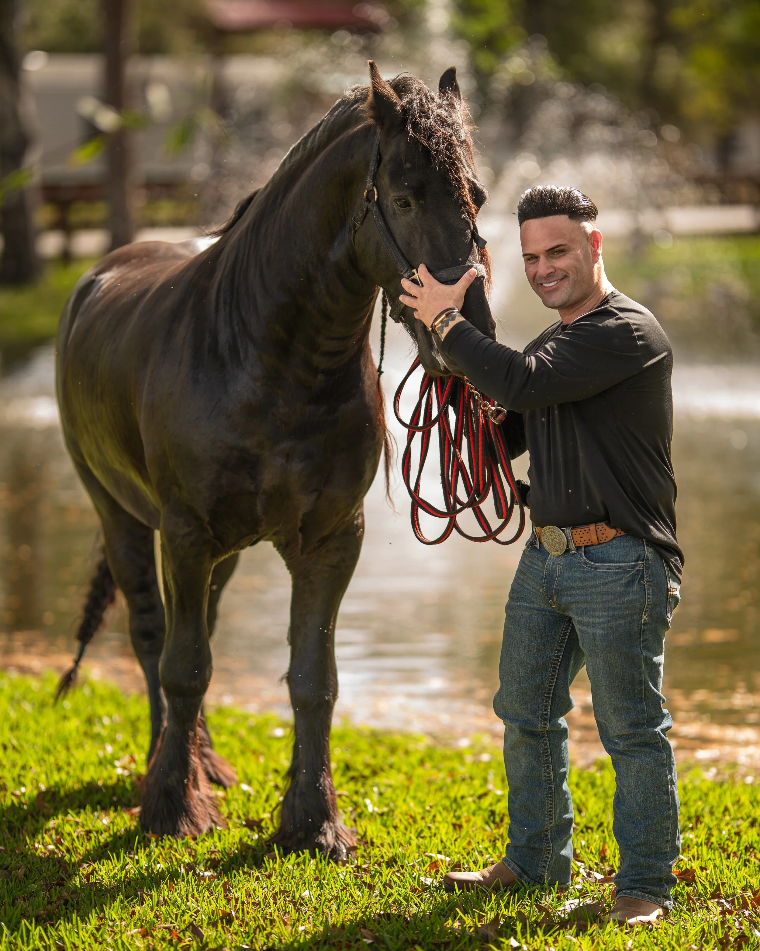 Luis Aquino  sonriendo sostenido la cabeza de un caballo negro junto a un río en un día soleado Vaqueros Del Pantano.
