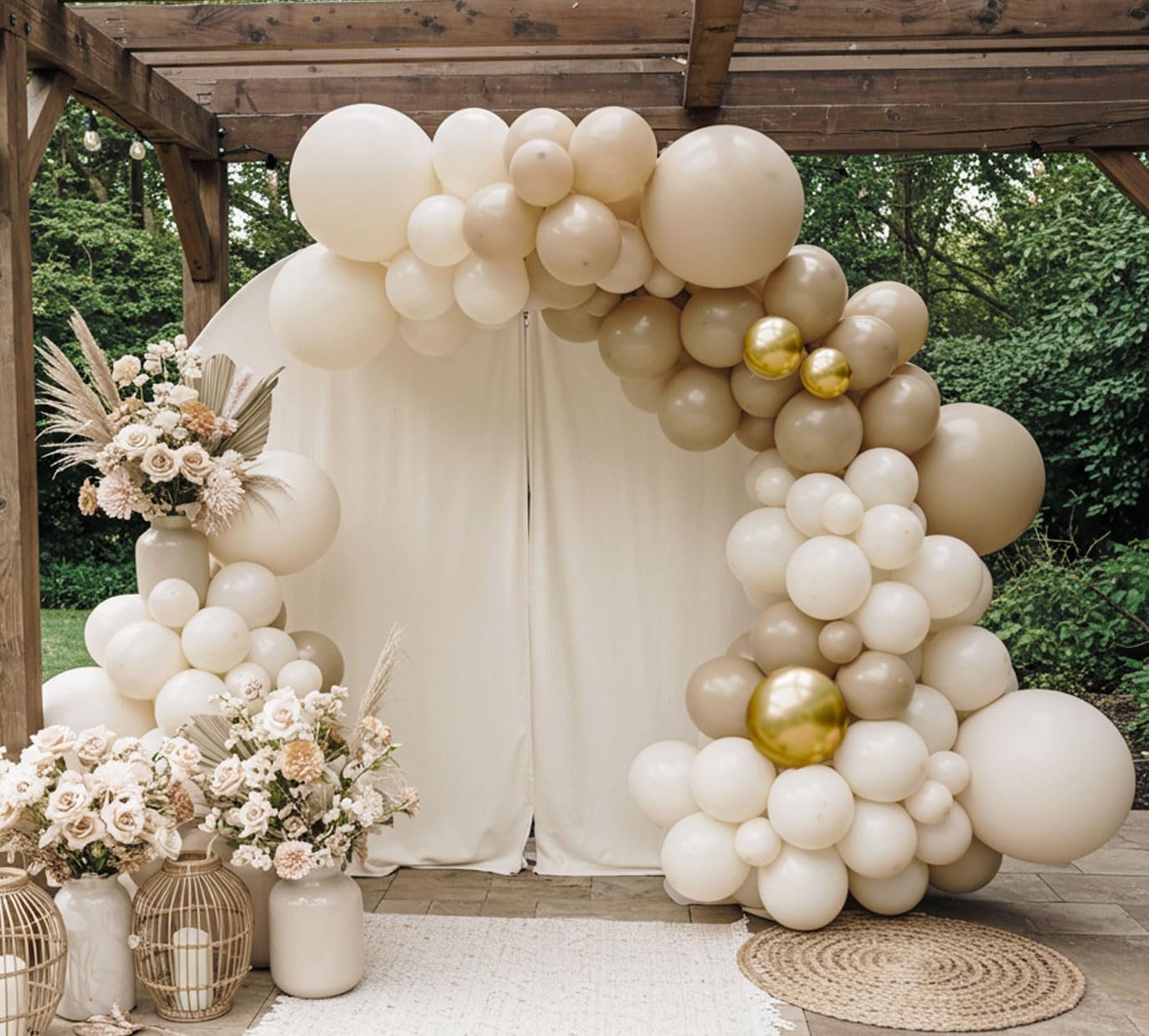 Decorative arch made of white, beige, and gold balloons, with floral arrangements in vases and wicker lanterns featuring candles, set in an outdoor space with greenery.