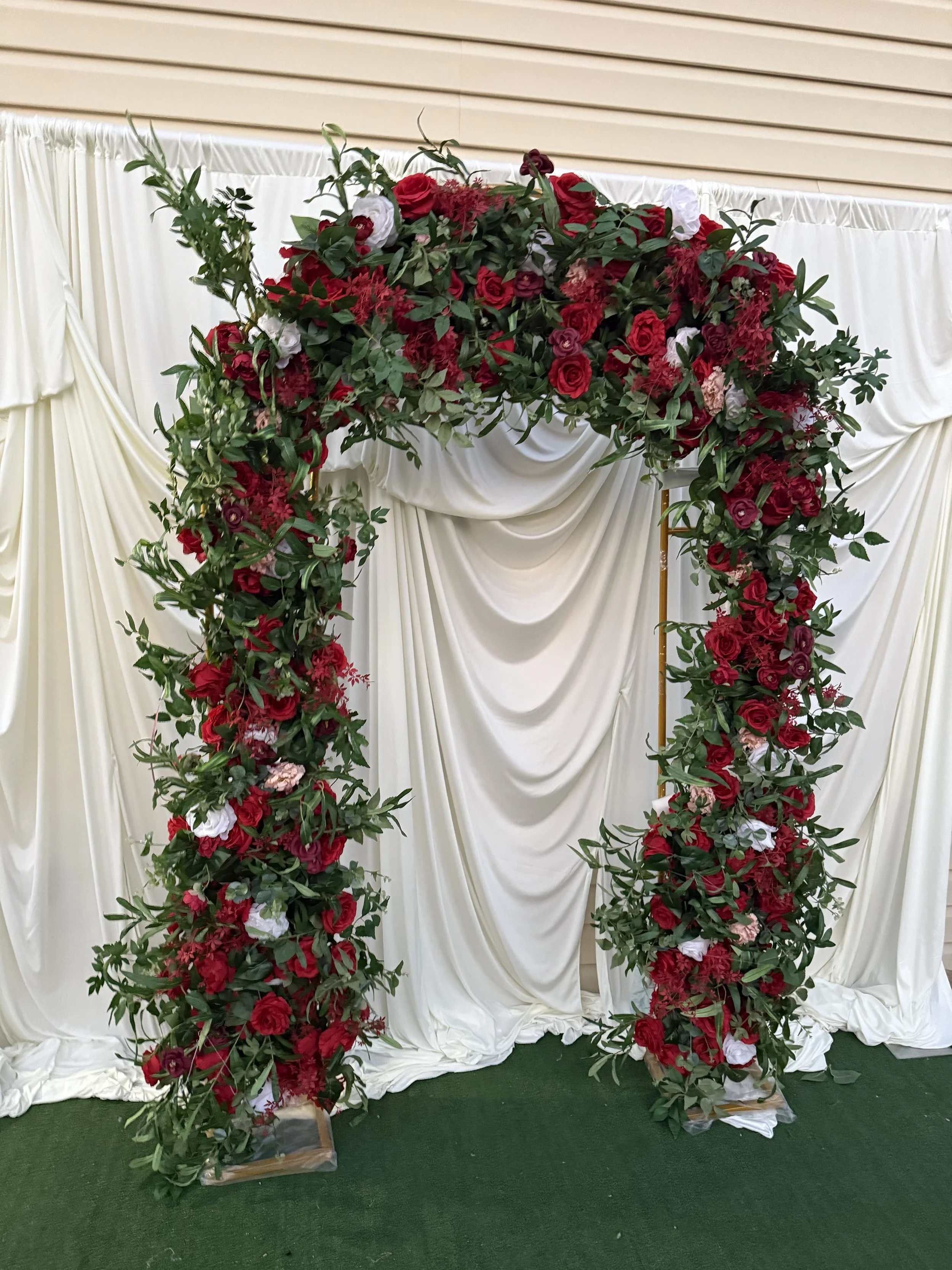 A decorated wedding arch with red and white flowers, green foliage, and a cream-colored fabric backdrop on a green carpet.