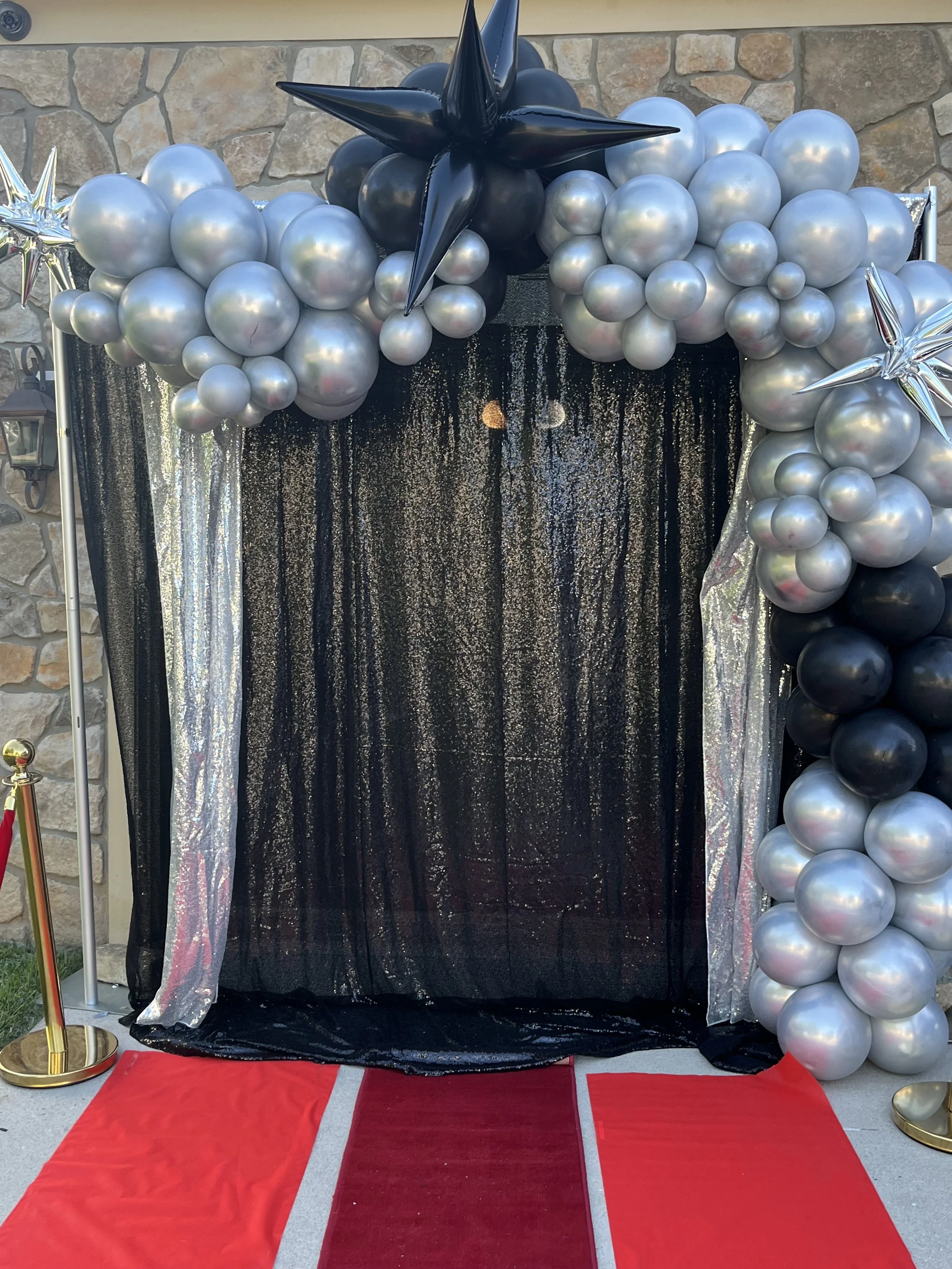 Black curtain with silver, black, and iridescent balloons arranged in an arch, decorated with large star-shaped balloons on the sides, a red carpet on the ground, and gold stanchions with red velvet ropes.