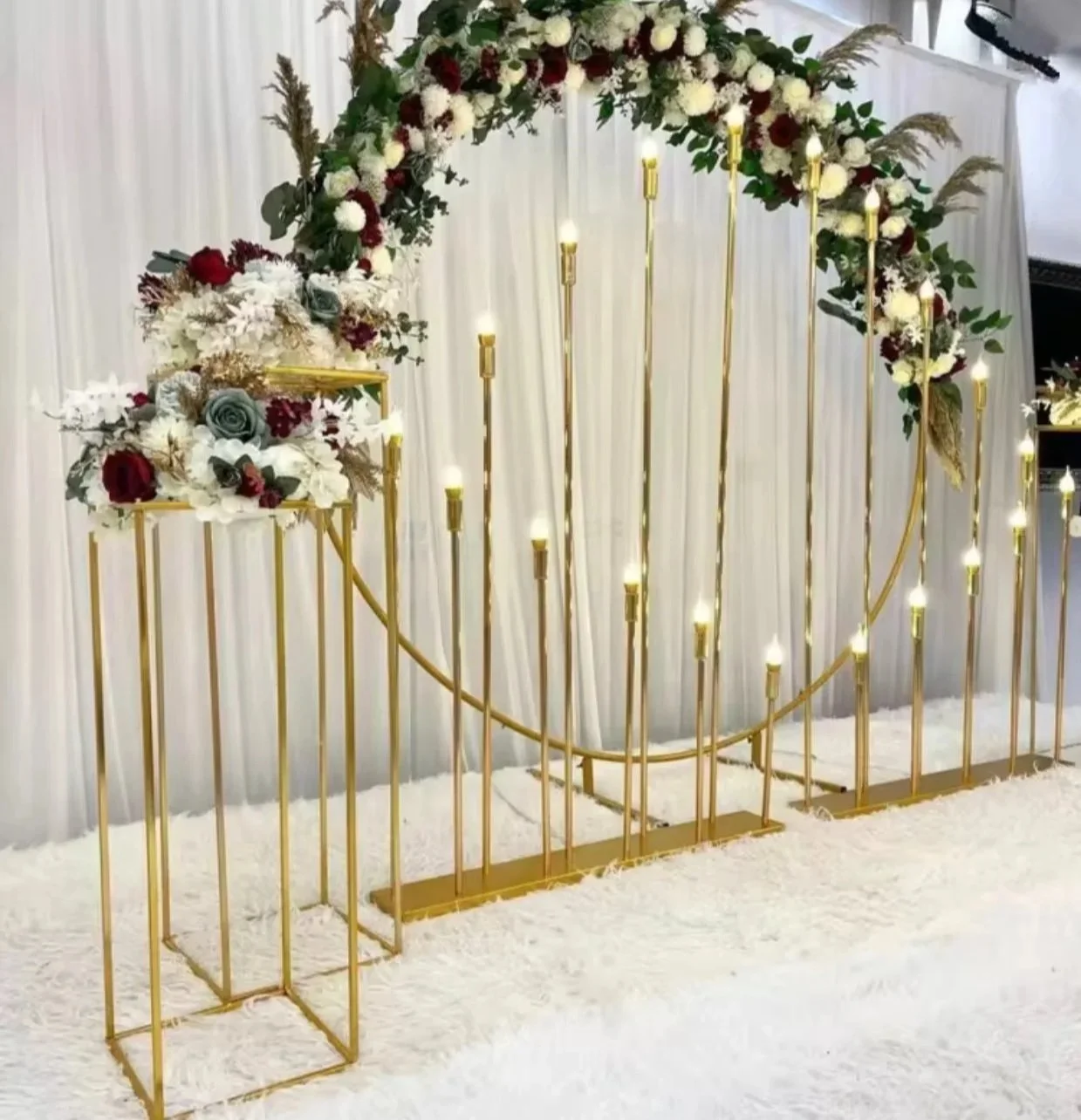 A decorative backdrop with a gold metal frame, floral arrangements, and LED candle lights, set on a white fluffy carpet in front of white curtains.