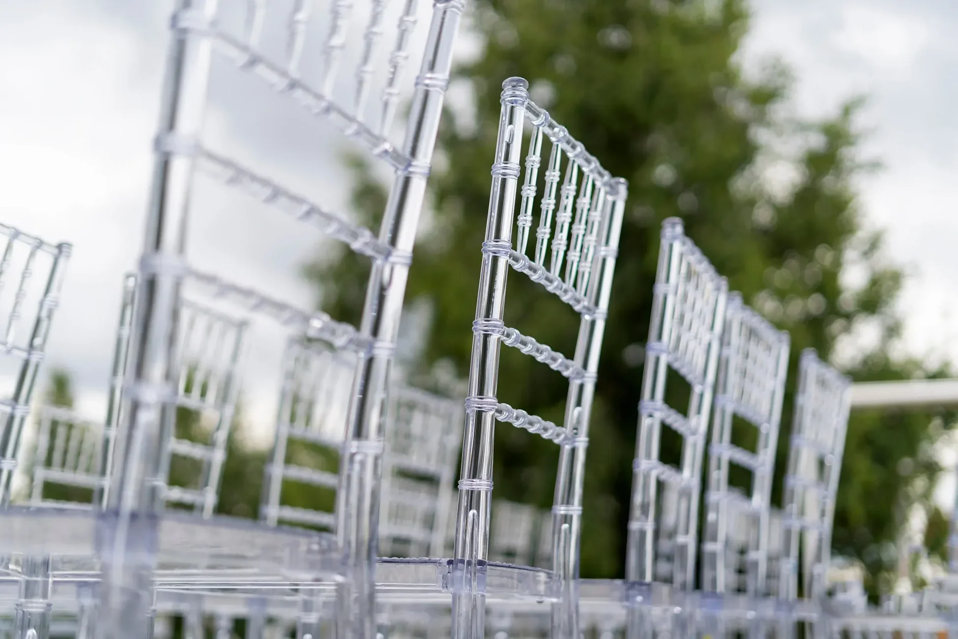 Close-up of empty transparent acrylic chairs outdoors with trees and cloudy sky in background.