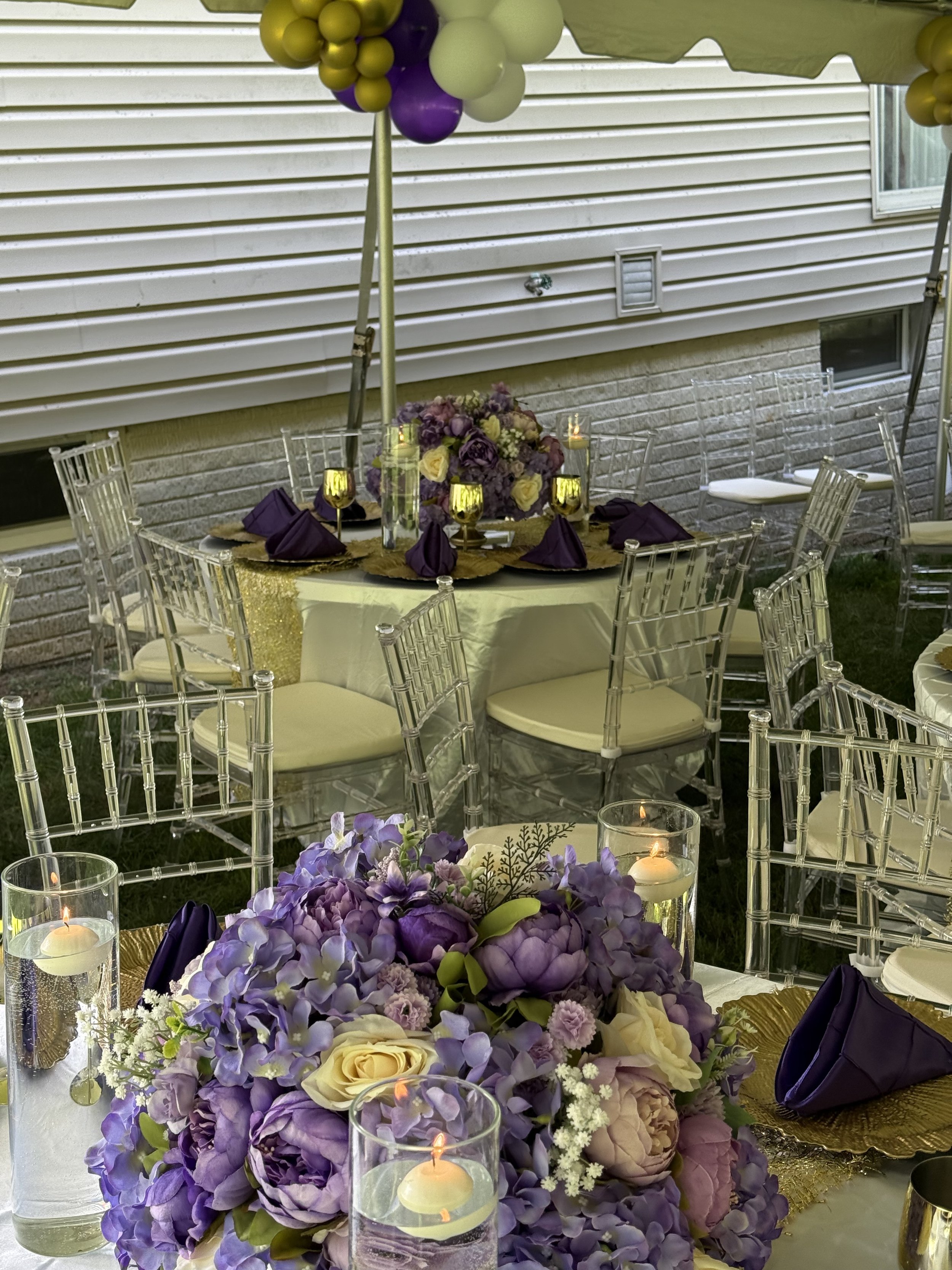 Elegant outdoor event setup with a round table decorated with purple and white floral arrangements, candles, and purple napkins, under a canopy with purple, white, and gold balloons, against a house with beige siding and brick foundation.