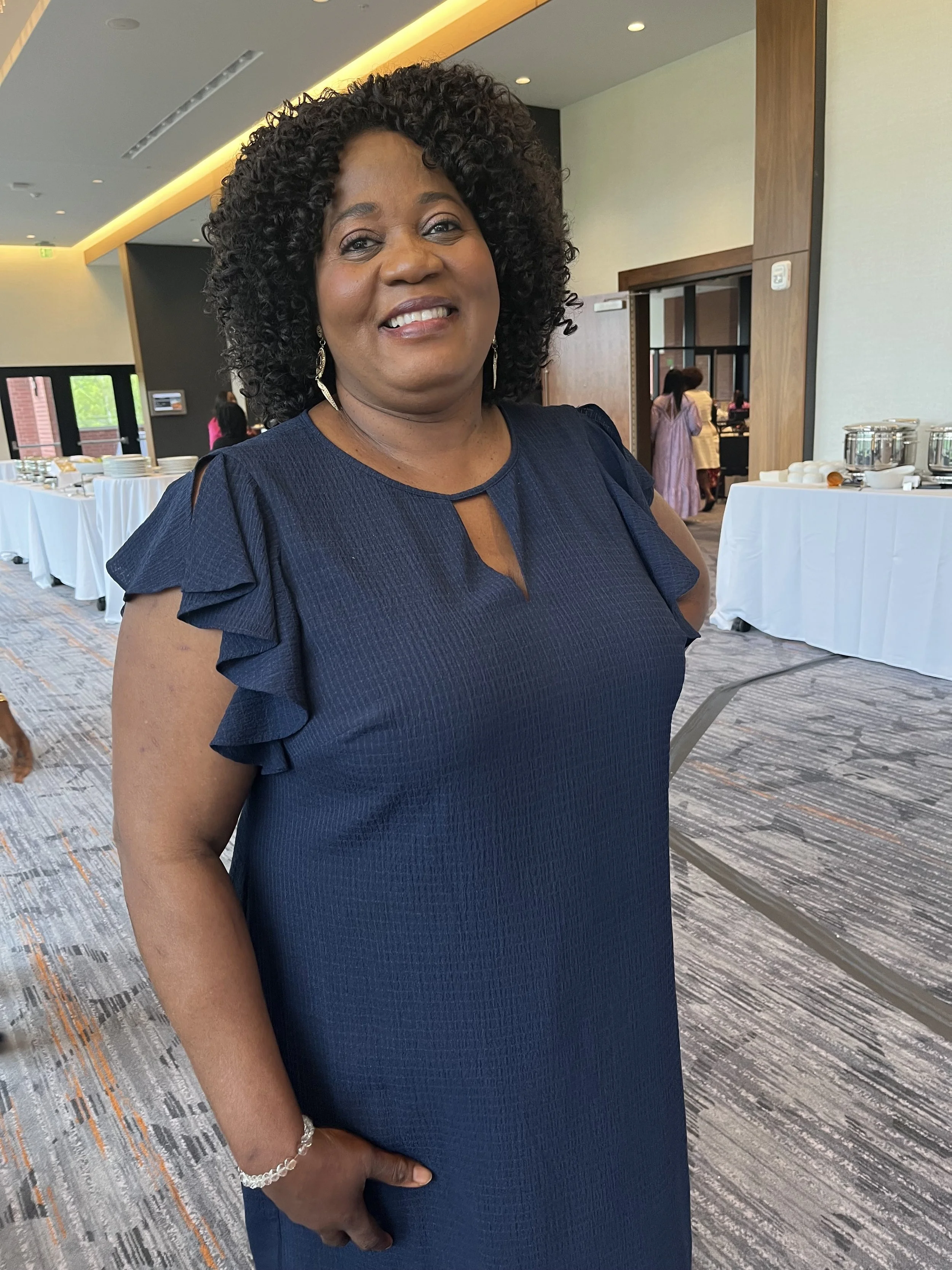 A woman in a navy blue dress with ruffled sleeves smiling at the camera in a banquet hall.