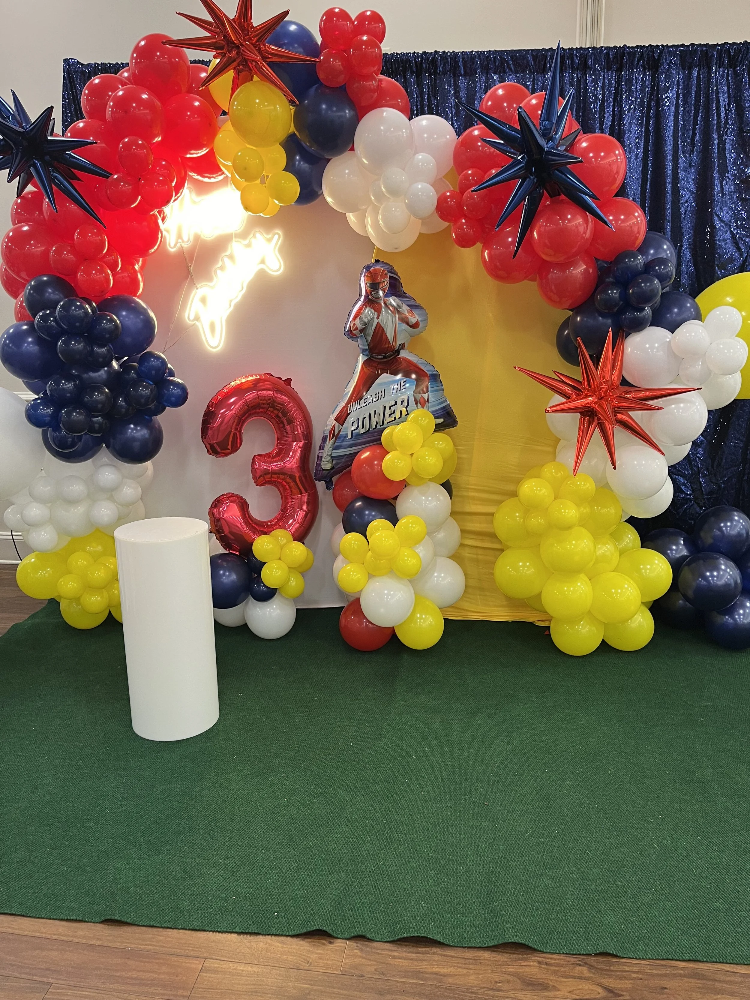 Colorful balloon display with a red number 3, a Power Ranger balloon, and star-shaped balloons in red and blue. Blue and white curtains in the background, and a green carpet on the floor.