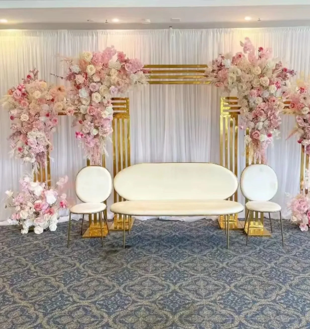 Floral wedding backdrop with pink and white flowers, gold frame, and beige seating arrangements on patterned carpet.