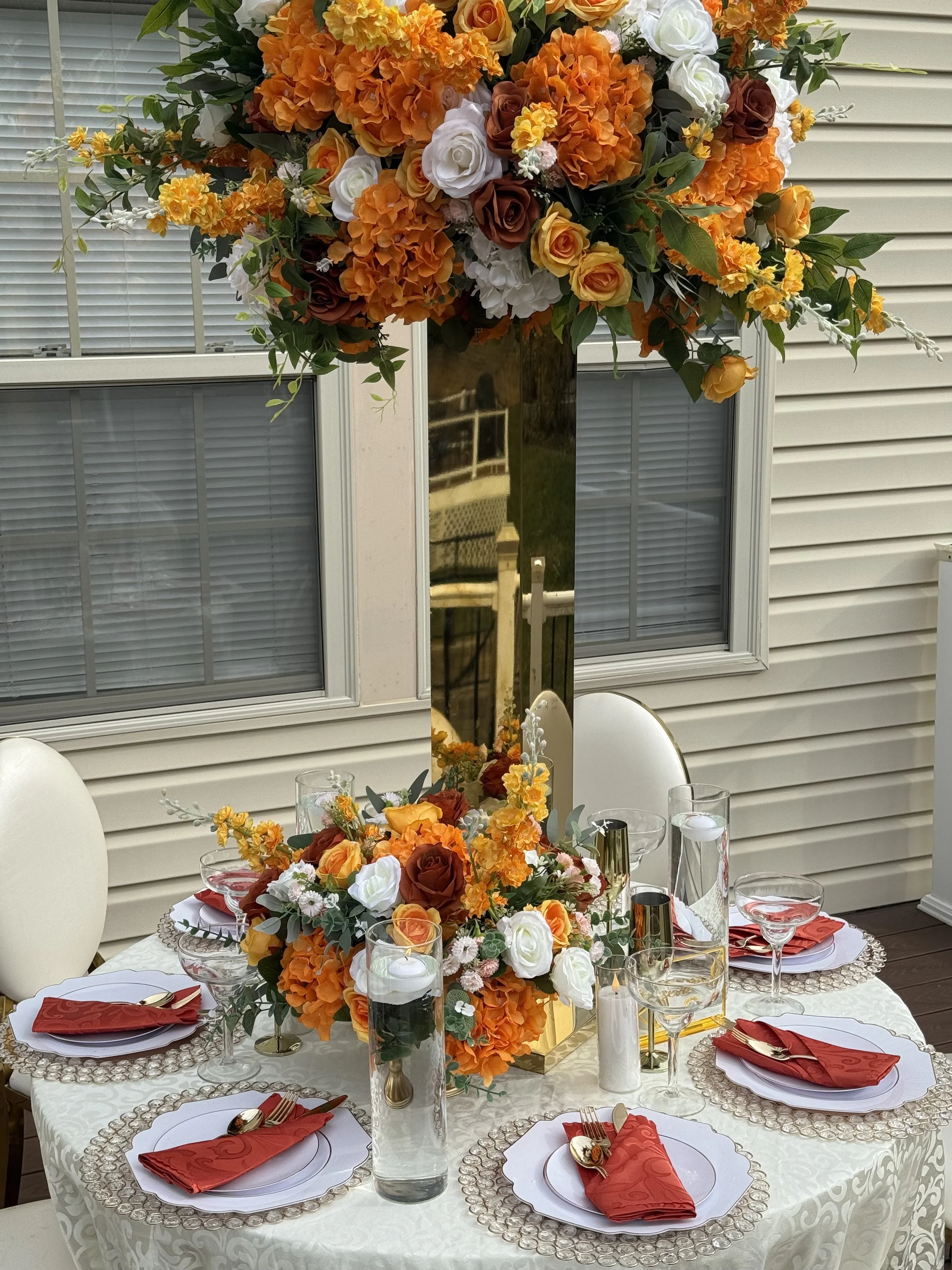 A decorated outdoor dining table with a tall floral centerpiece featuring orange, white, and yellow flowers, set for six people with white plates, red napkins, gold utensils, and glassware, with a mirror reflecting the floral arrangement.