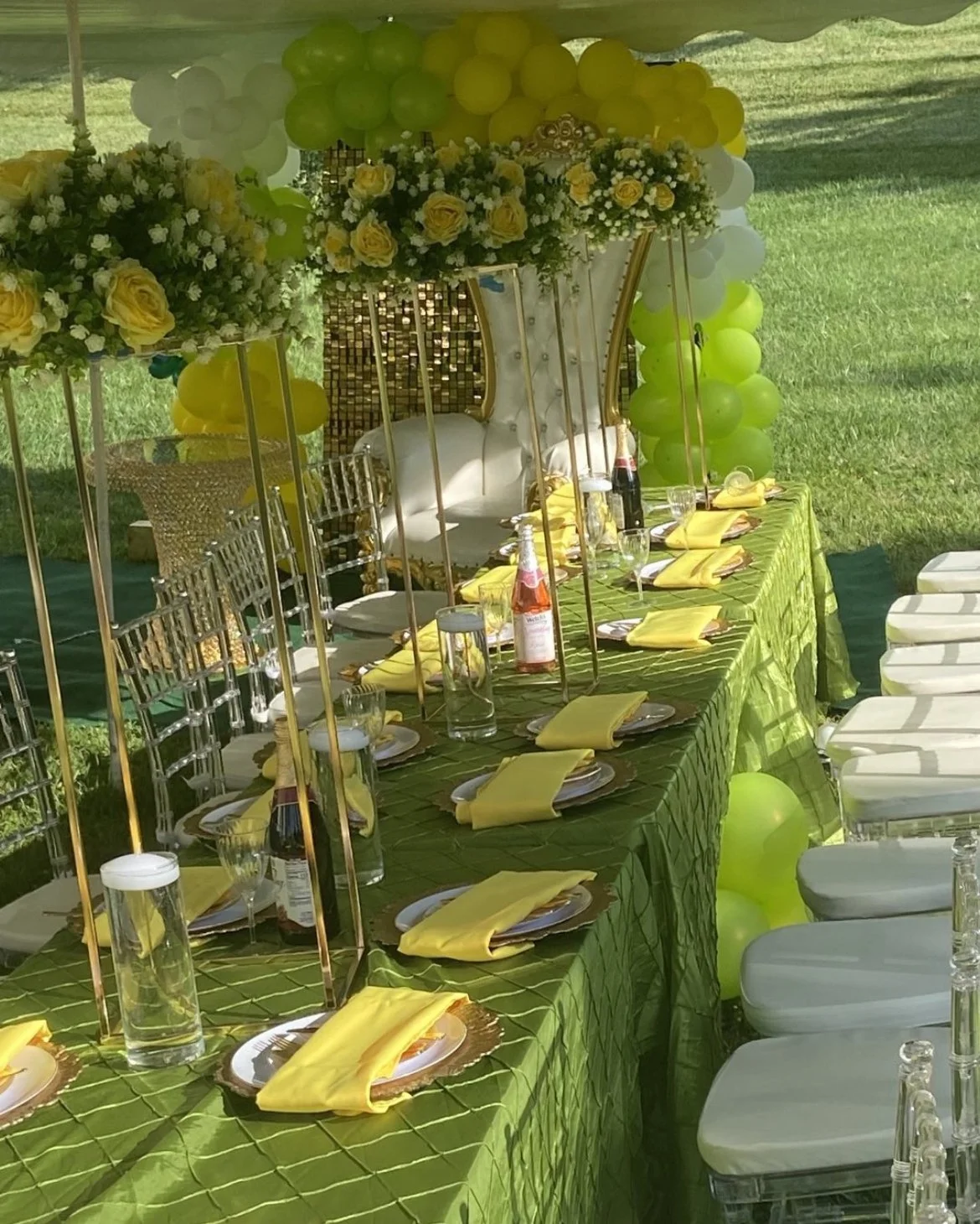 Table set up outdoors with yellow napkins, glassware, and bottles, decorated with yellow and white flowers and balloons, featuring a high-backed white and gold chair at one end.