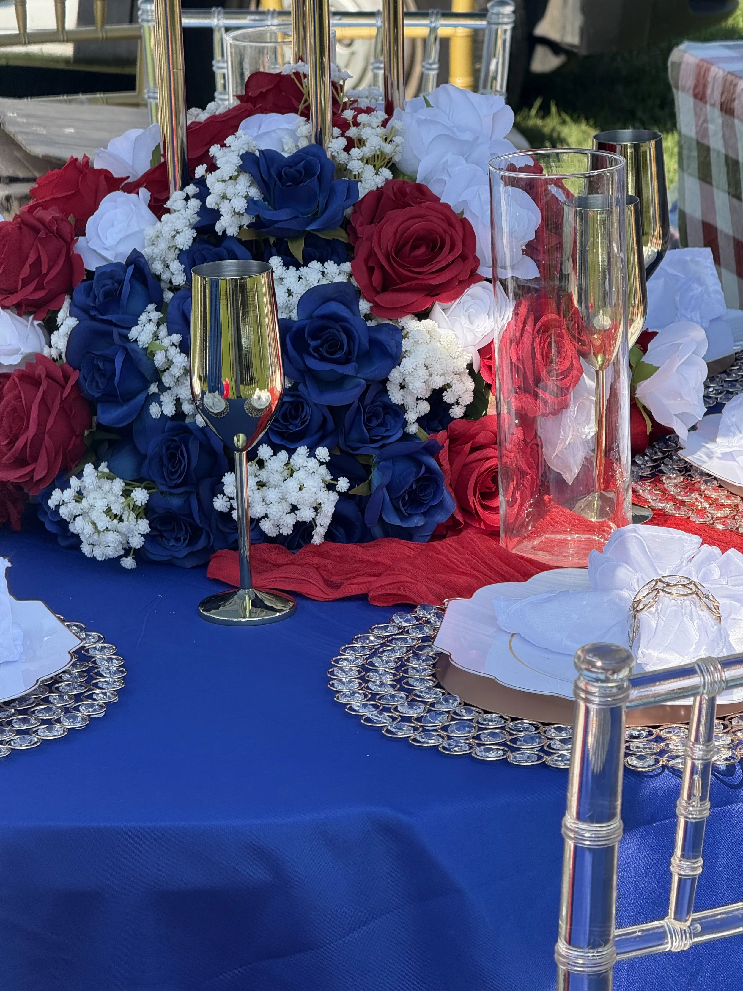 A decorated table with a large centerpiece of red, white, and blue roses, surrounded by champagne glasses, a tall glass vase, and place settings with white napkins and decorative chargers, set outdoors with sunlight on a checkered chair in the background.