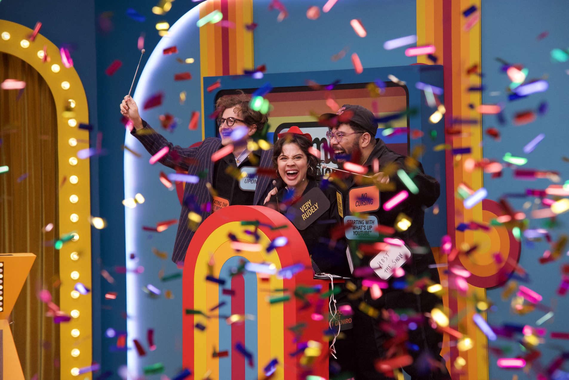 Three people celebrating with colorful confetti, standing behind a game show podium with a rainbow target design, surrounded by bright yellow and orange decorations, smiling and laughing