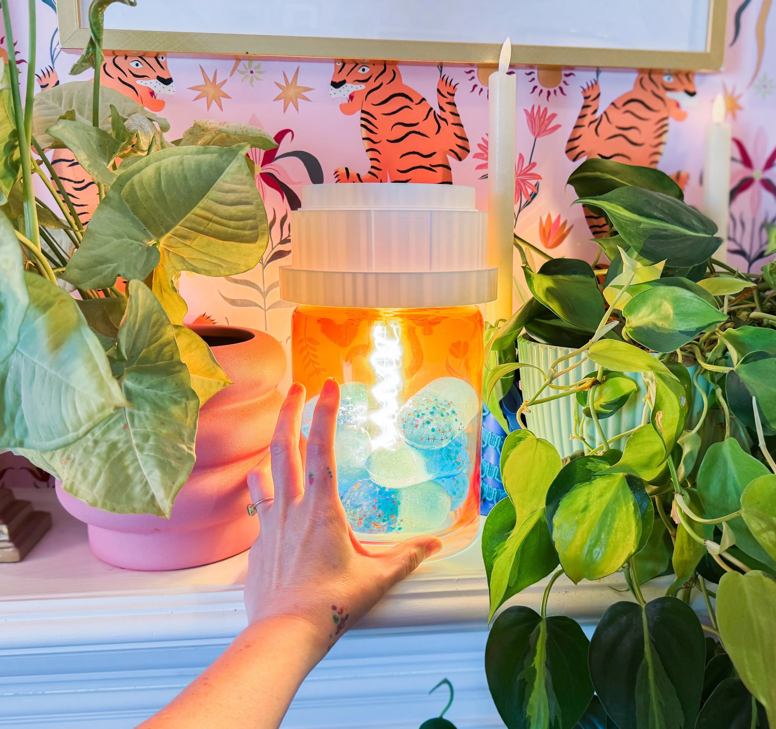 Person holding a giant pill bottle lamp glass vase filled with illuminated fairy lights behind green houseplants, with a pink and tiger-themed background.