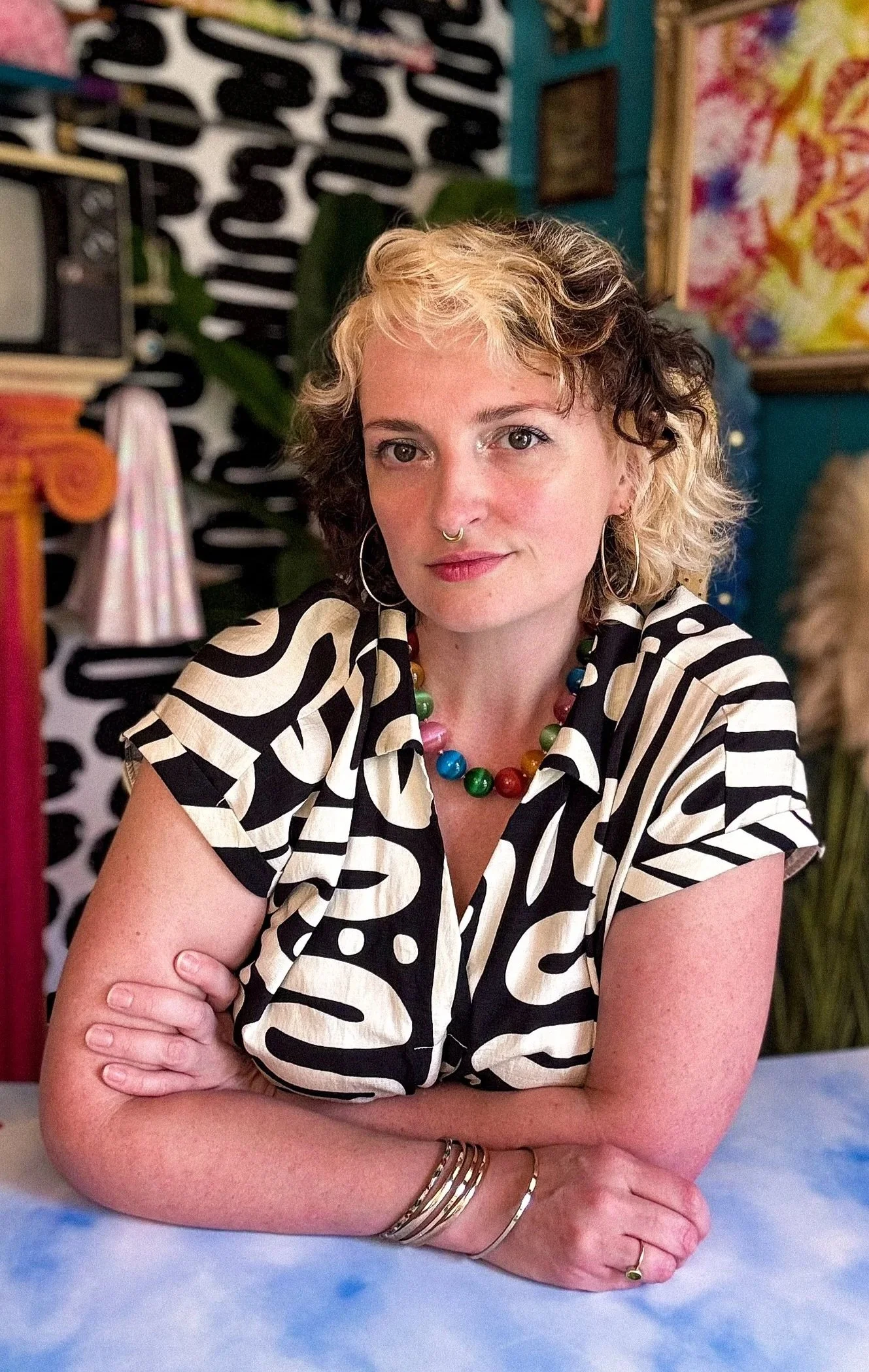 A woman with short, curly, blonde and brown hair, wearing a black and white abstract patterned shirt, a colorful beaded necklace, and hoop earrings. She has a septum piercing and is sitting at a table with her arms crossed in front. The background features art pieces and colorful decor.