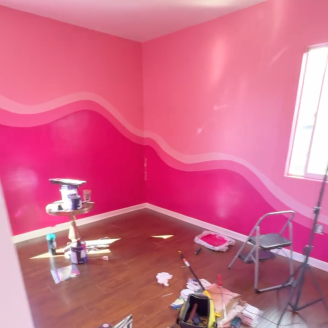 Room with pink walls featuring a wavy mural, wooden floor, small window, and painting supplies scattered around.