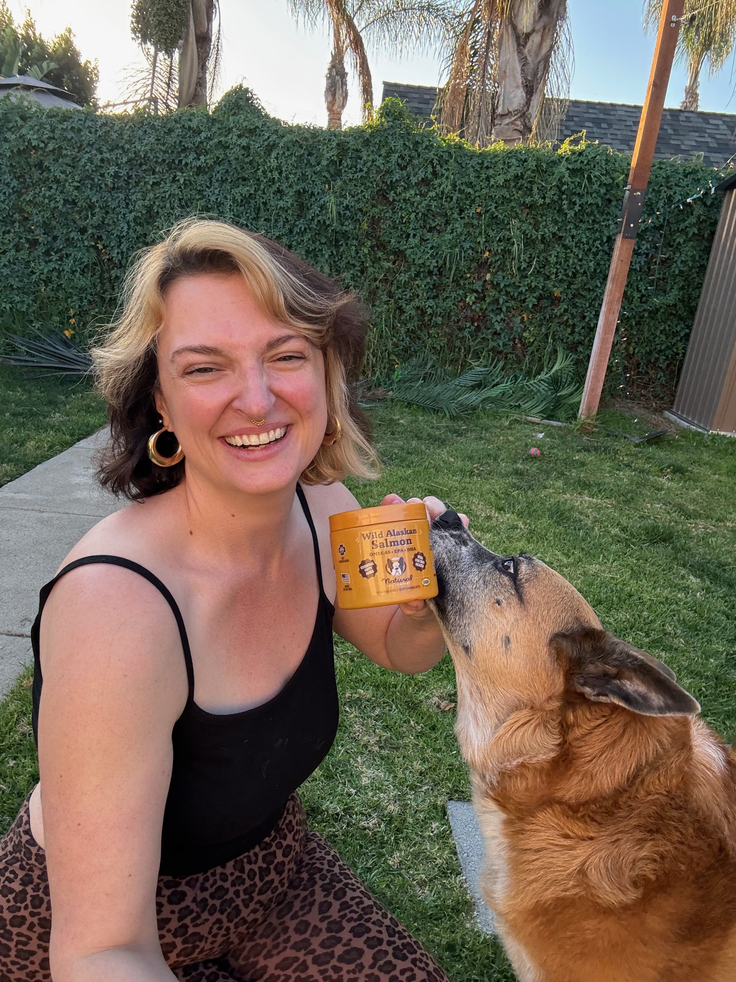 A woman with short, wavy blonde and brown hair, wearing a black tank top and leopard print pants, smiling while holding a container of Wild Alaskan Salmon dog food. She is outdoors on a grass lawn, with a large brown dog licking her face. The background features green hedge, trees, and a wooden and metal structure.