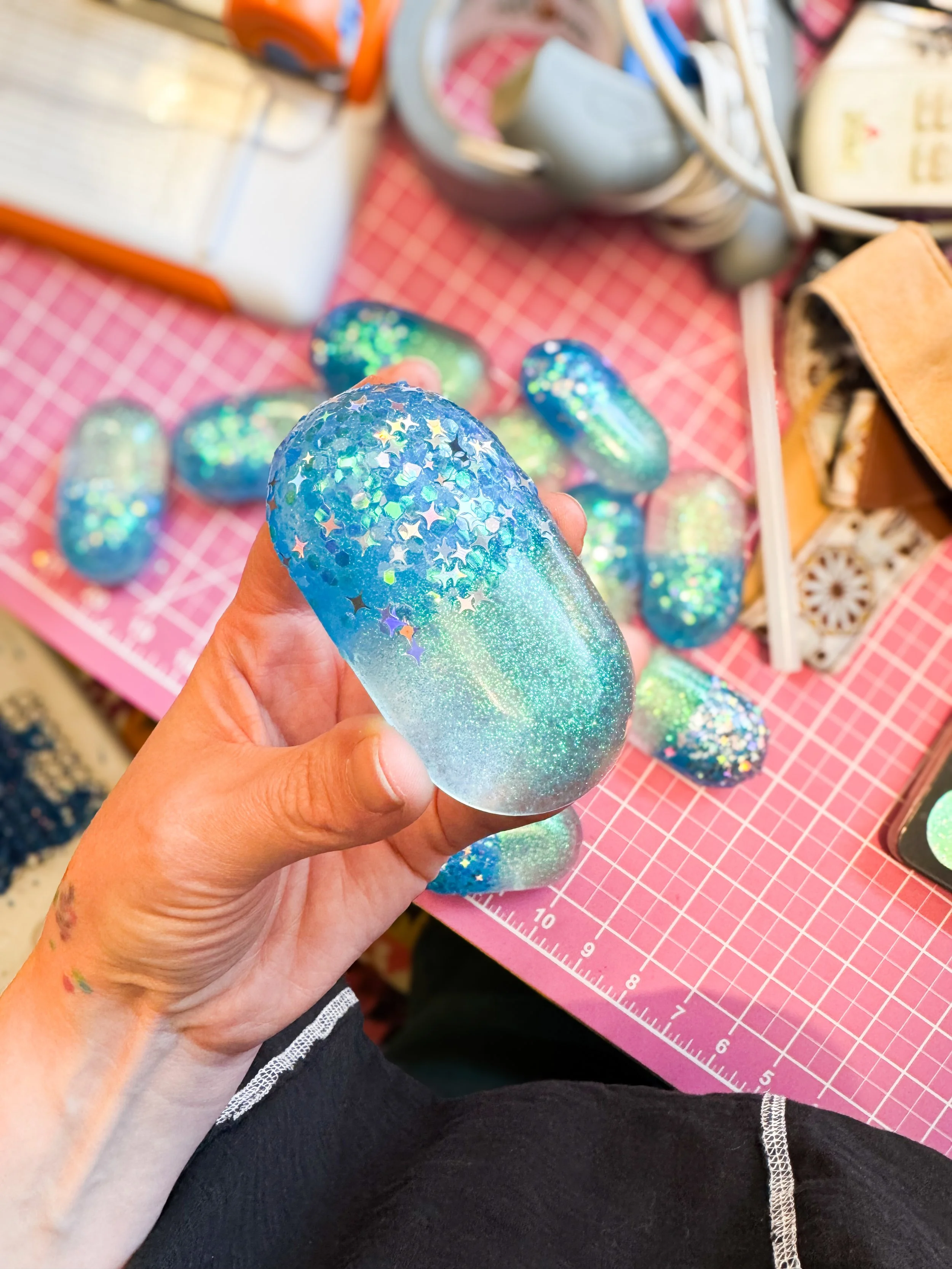 A person holding a holographic, iridescent, decorative glass stone with star-shaped glitter in front of a pink grid craft mat, with several similar stones on the mat, and hobby supplies and tools in the background.