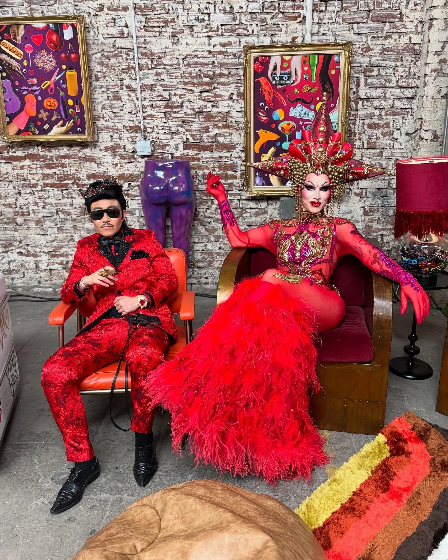 A man in a red patterned suit with sunglasses and a woman in elaborate red and gold costume with a large headdress, sitting in an industrial-style room with a brick wall, colorful artwork, and eclectic furniture.