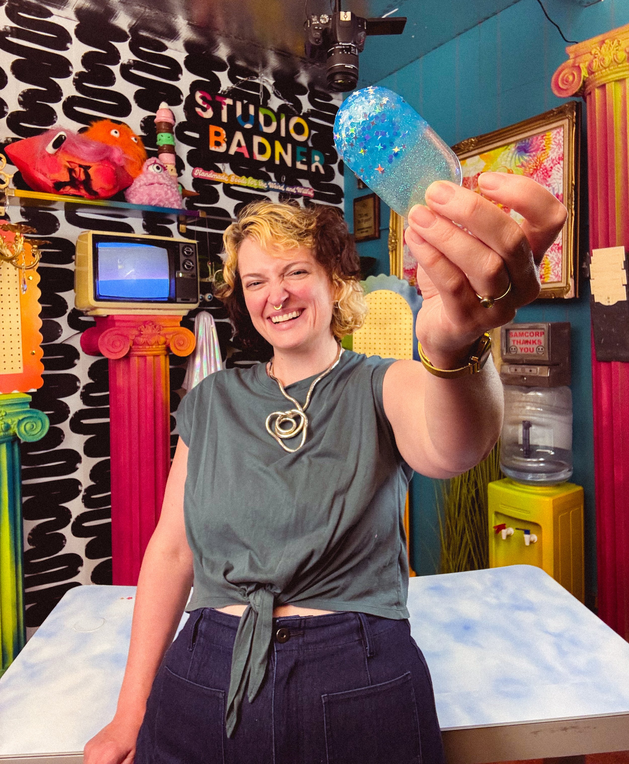 A person holding a glittery, colorful acrylic resin pill in front of a decorated and vibrant room with toys, artwork, and a sign that reads 'STUDIO BADNER'.